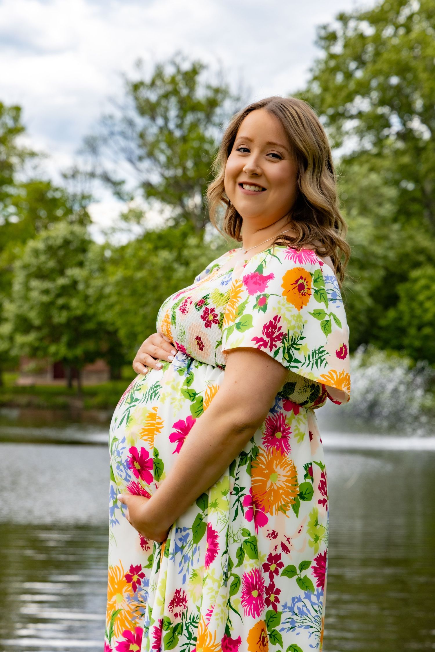 A person in a vibrant floral maternity dress stands by a pond with a fountain in the background.