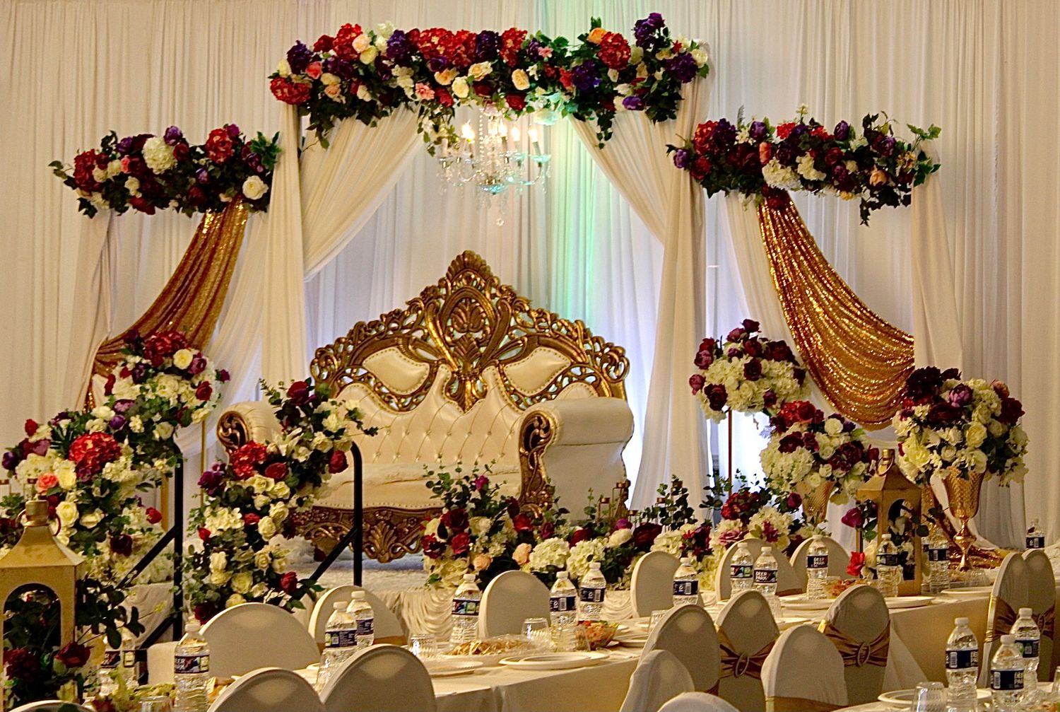 Elegant wedding reception setup with ornate gold arch, floral garlands, and white draping under warm lighting.