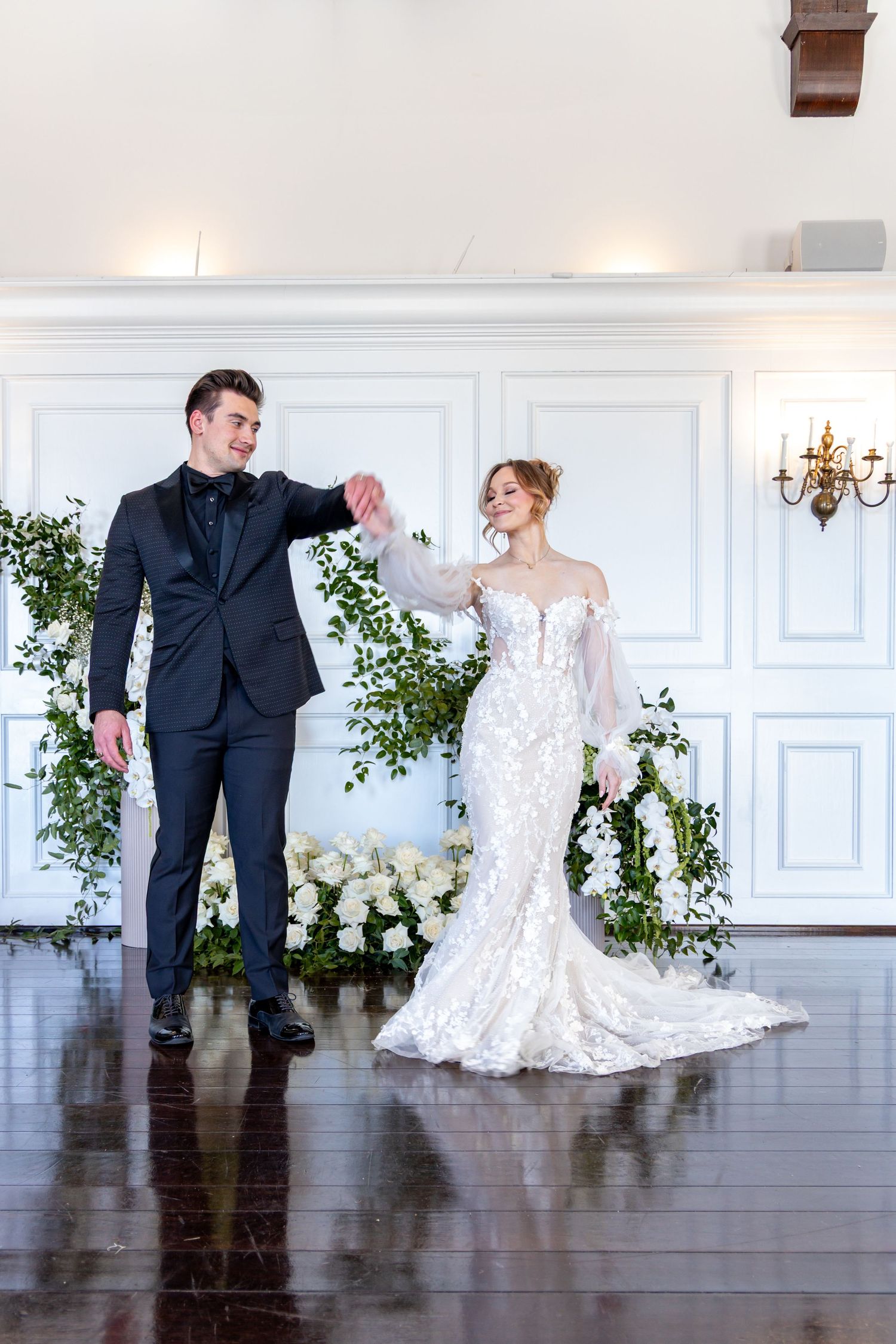 Wedding couple in formal attire poses in elegant white room with greenery and floral decorations.