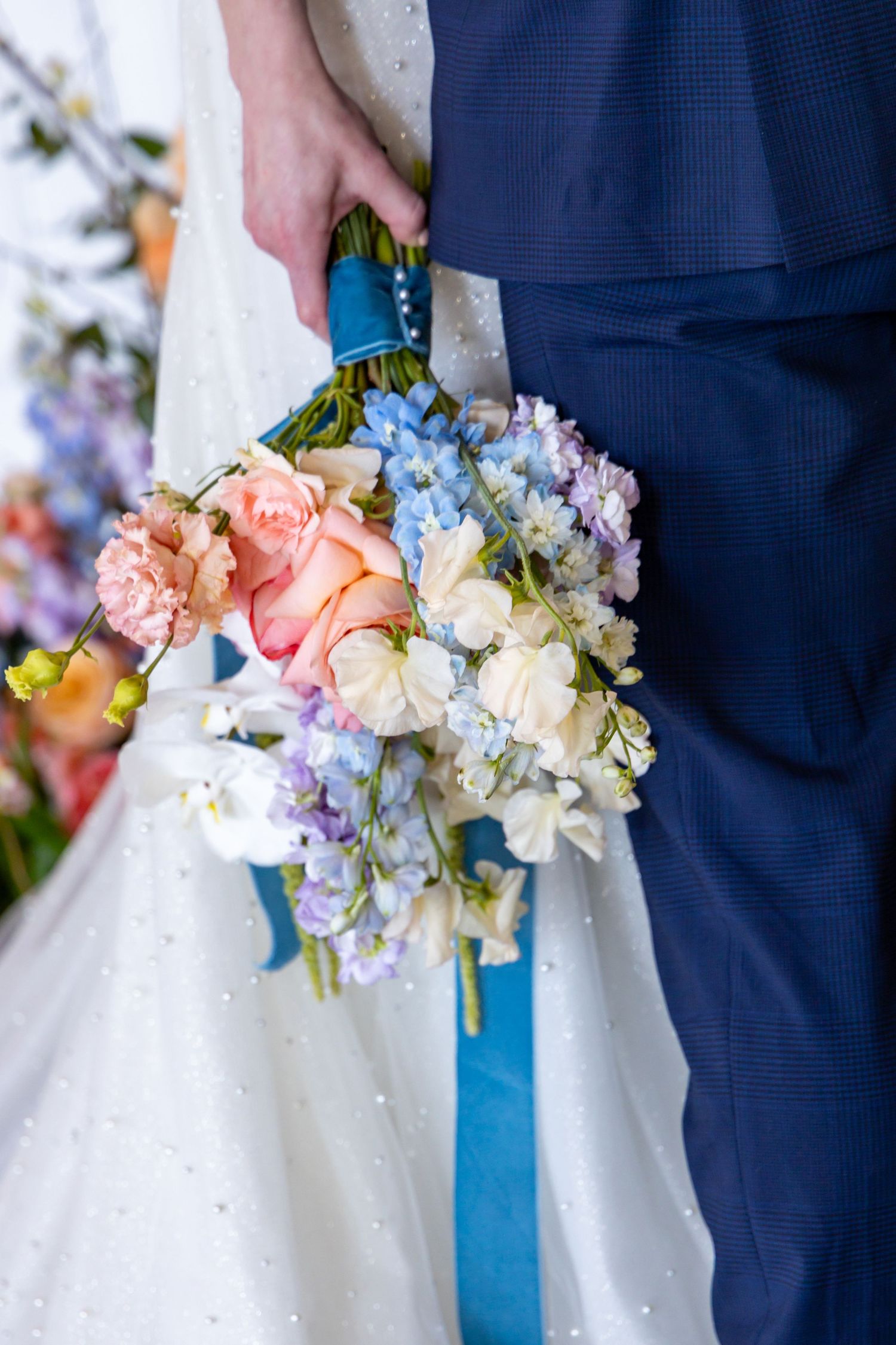 A wedding bouquet with pink and blue hydrangeas wrapped in turquoise ribbon held against a white dress and navy suit.