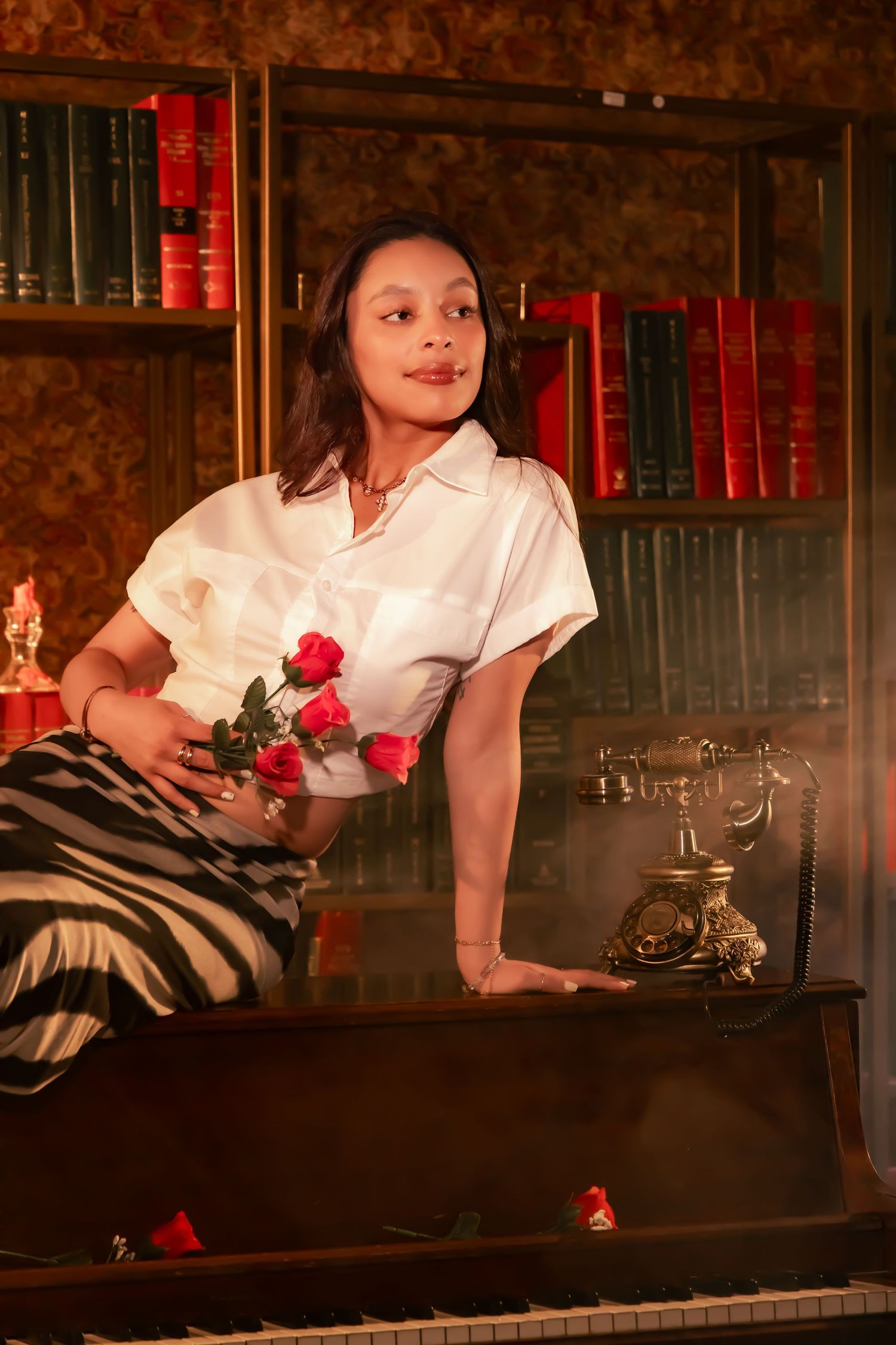 A figure in a white blouse poses with red roses by a vintage piano with antique books and a rotary phone in the background.