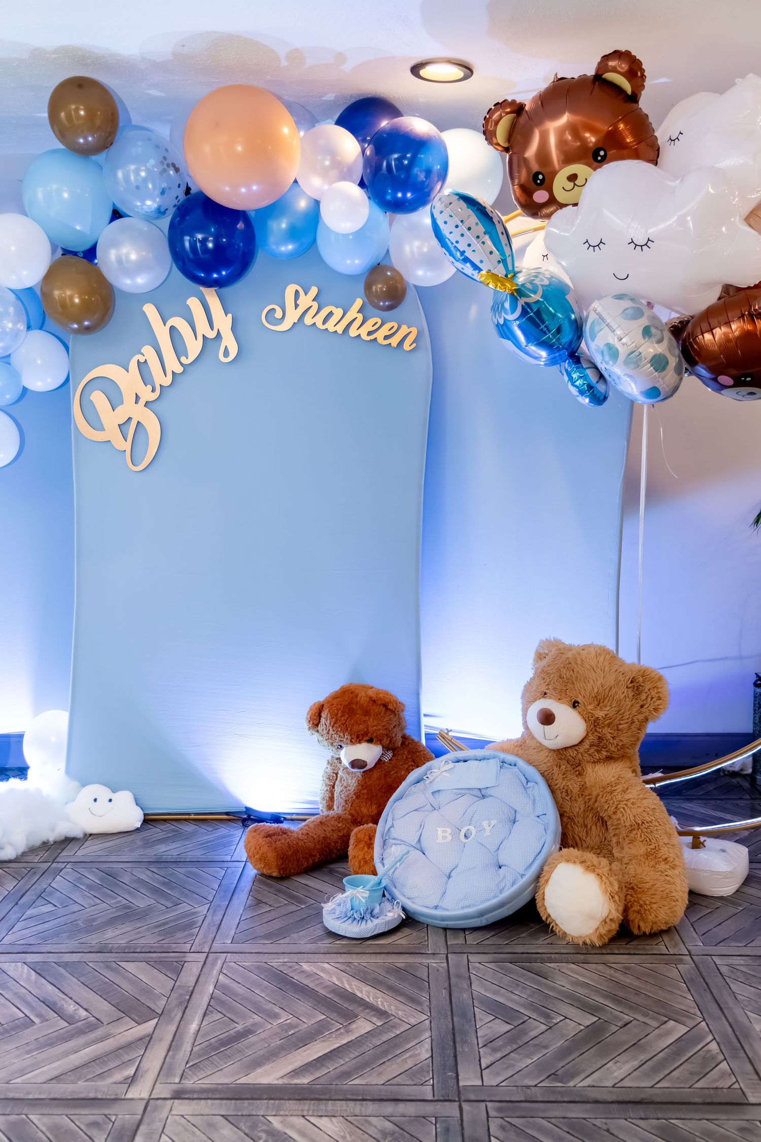 A baby shower setup with blue balloon arch, teddy bear decor, and cursive 'Baby' lettering against a blue backdrop.