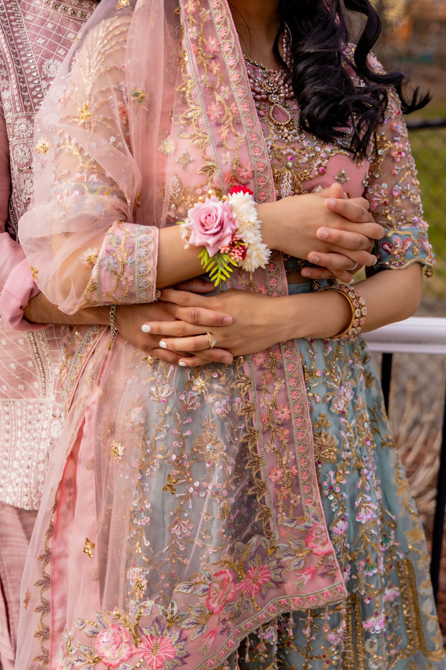 Close-up of hands wearing elegant jewelry and holding pink rose corsage against embroidered pink and blue floral lehenga.