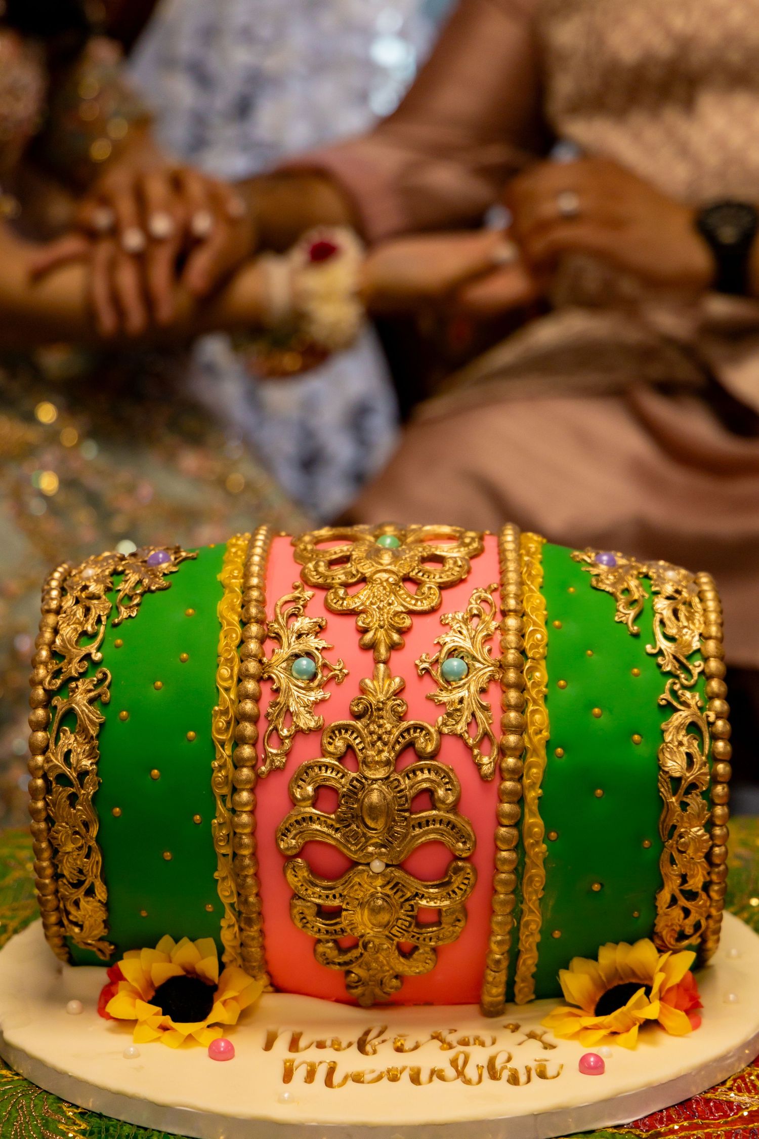 A luxurious Indian wedding cake decorated with green and pink fondant, gold details, and sunflower accents.