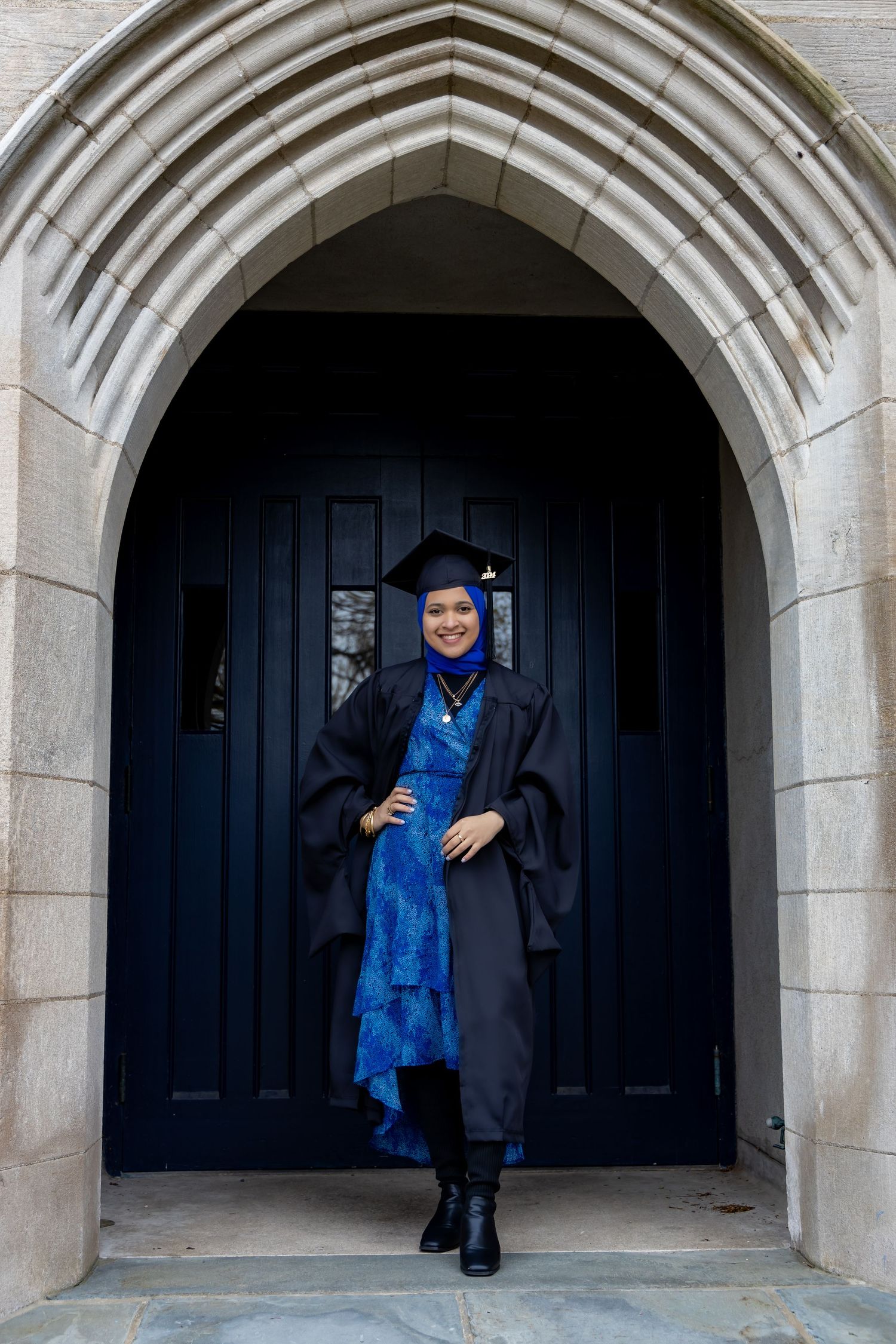 A graduate in academic regalia stands in a Gothic stone archway doorway wearing a blue dress and black boots.