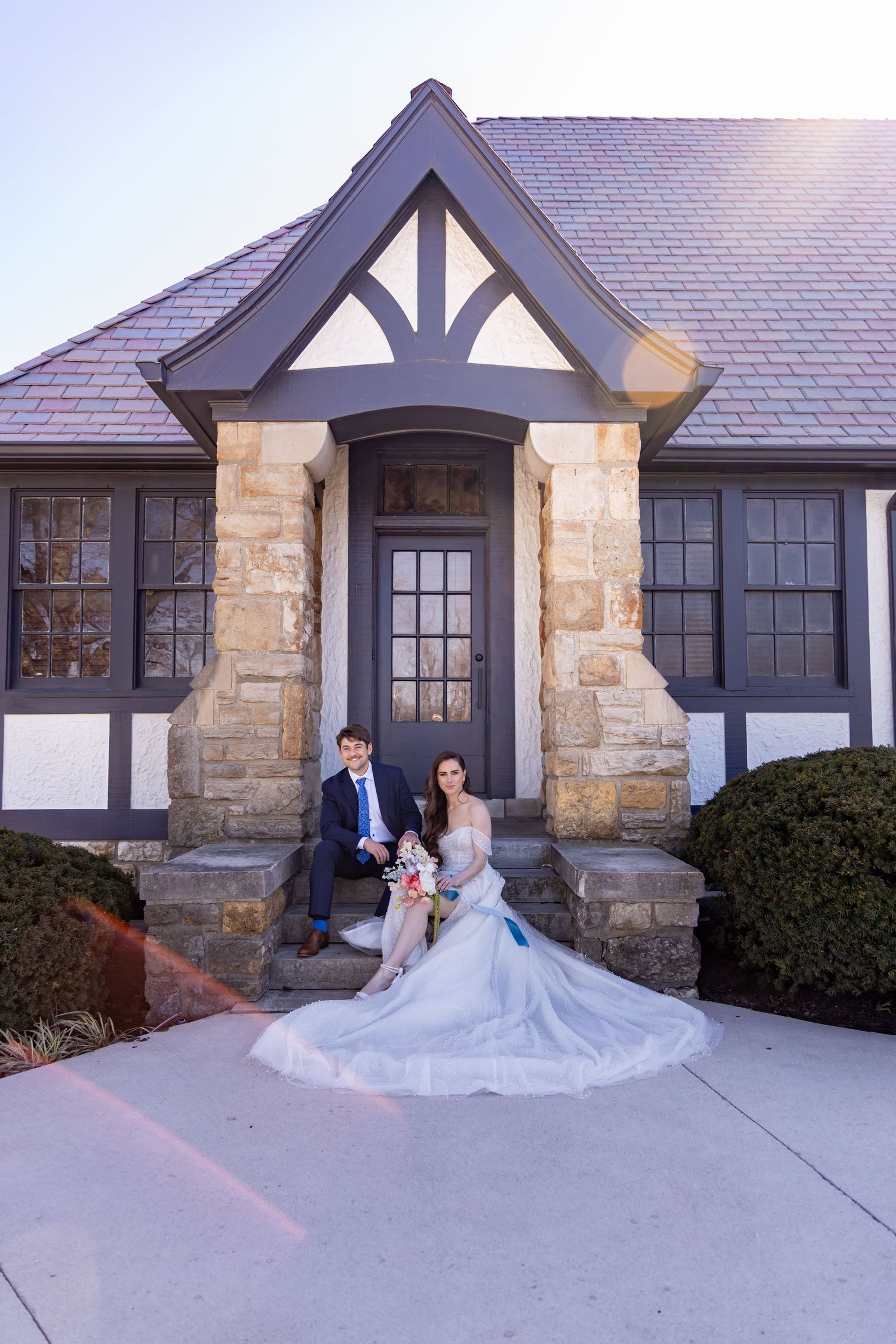 Elegant Tudor-style house with stone facade and gabled entrance featuring formal attire in front at sunset.