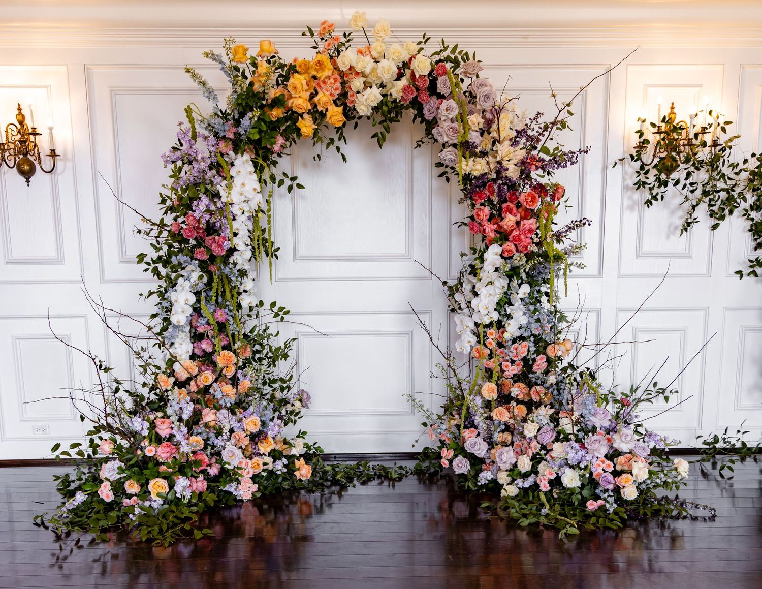 Stunning floral arch with colorful blooms and branches against white paneled wall with gold sconces.