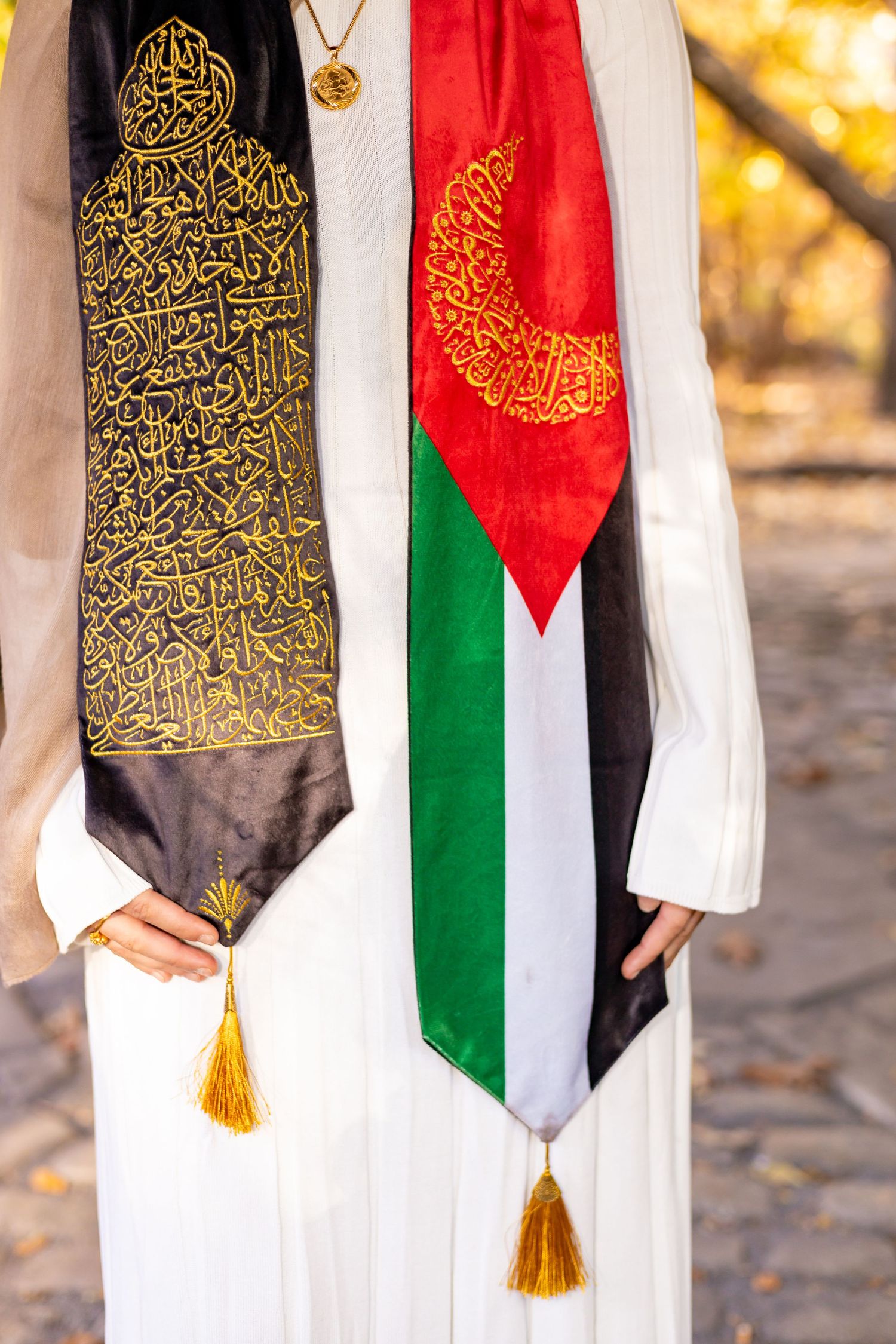Close-up of decorative Arabic calligraphy scarf and Palestinian flag scarf with gold tassels worn over white clothing.
