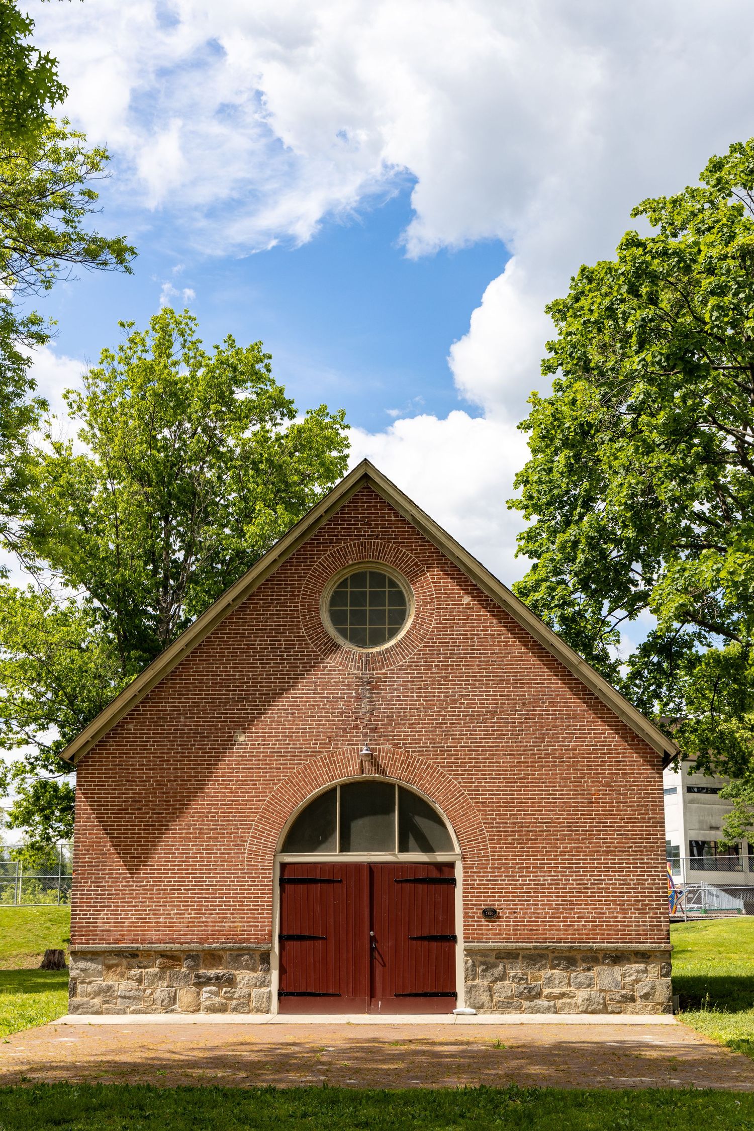 Small red brick chapel with wooden double doors and round window sits among green trees under a blue summer sky.