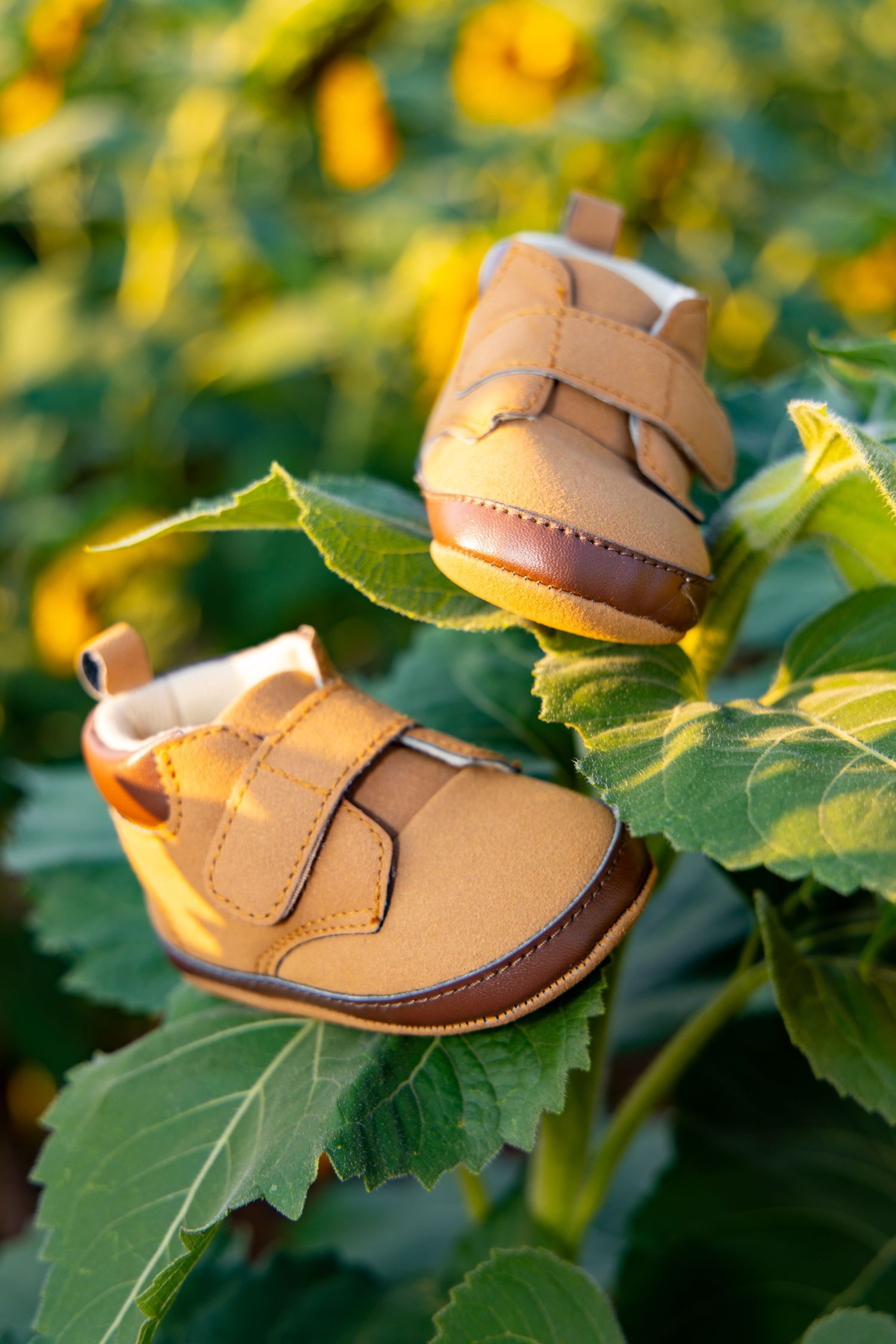 Tiny tan leather baby booties with velcro straps rest on green leaves against a blurred yellow floral background.