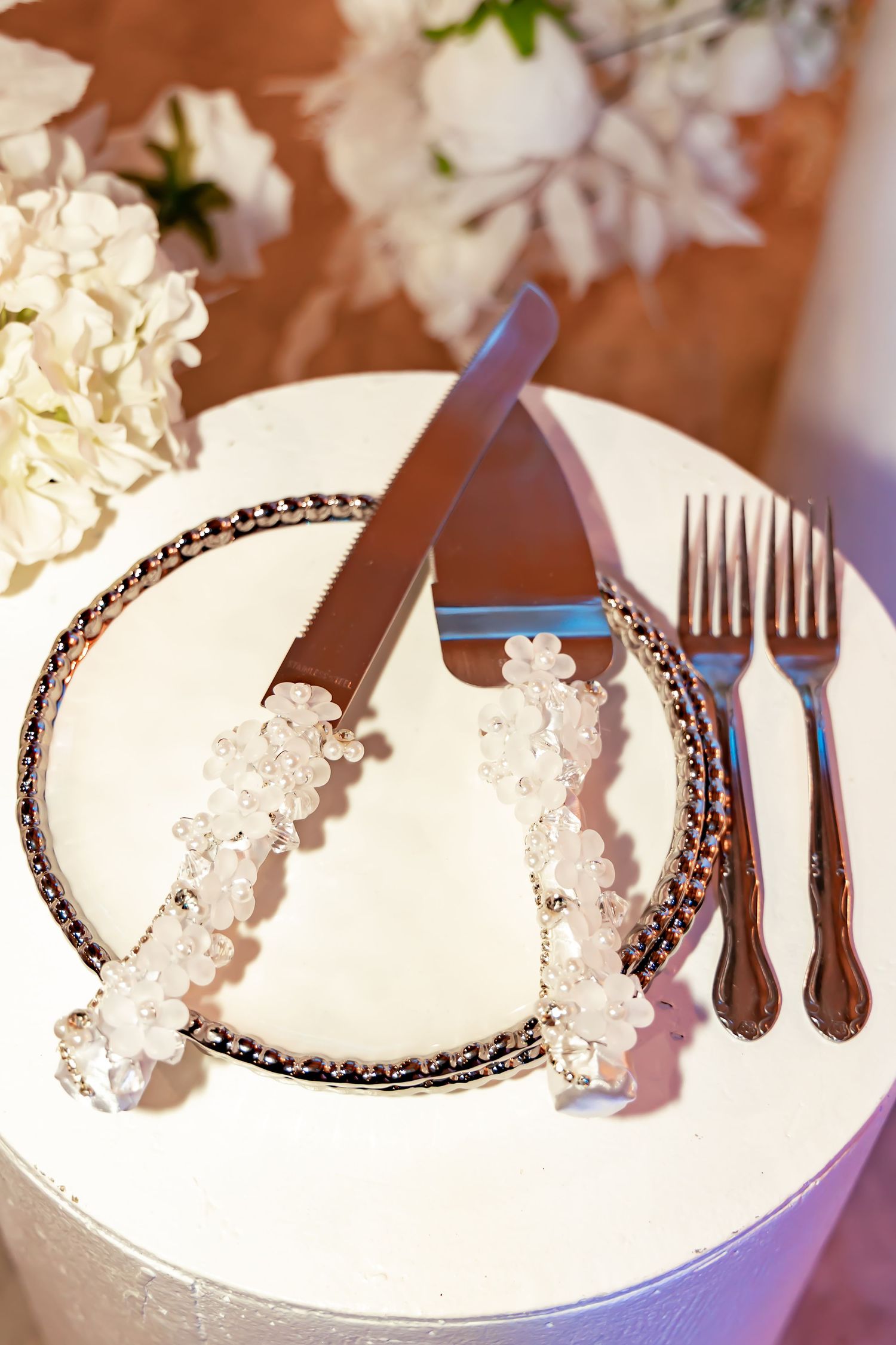 Elegant place setting with decorated utensils and beaded plate rim against white surface with flowers in background.