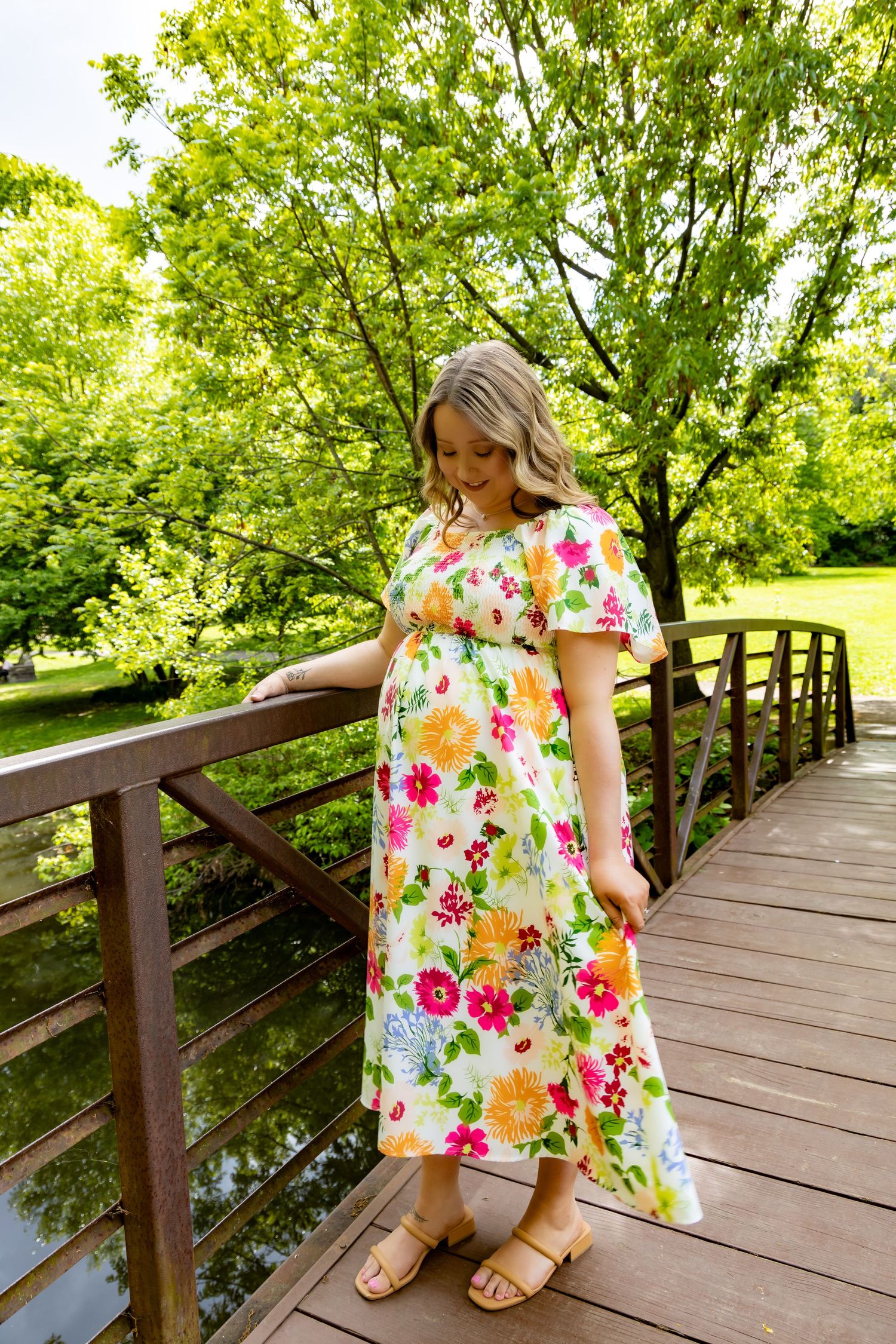 Someone wearing a colorful floral print maxi dress stands on a wooden bridge walkway surrounded by lush green trees.
