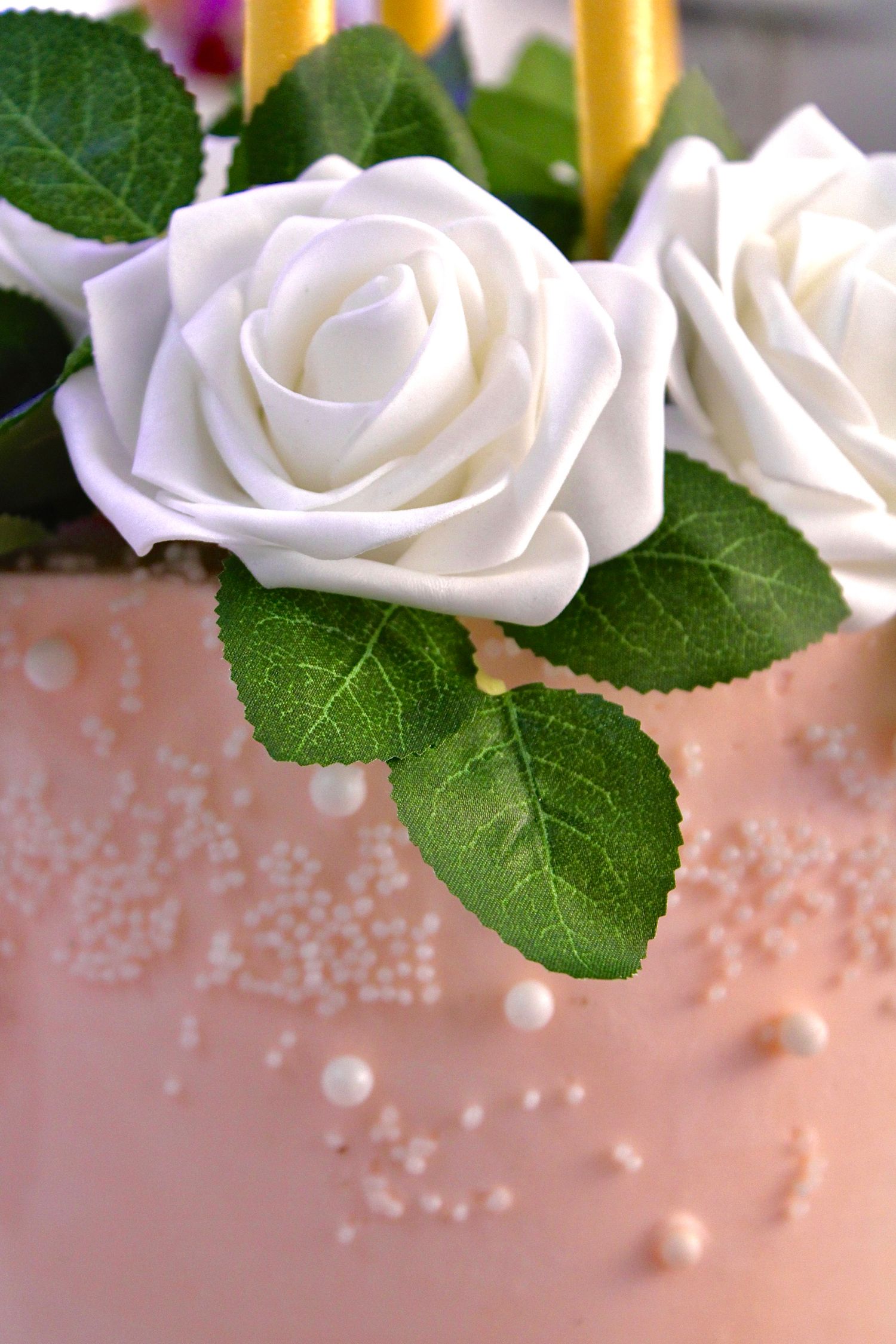White fondant roses with green leaves adorn a pink frosted cake decorated with delicate pearl accents.