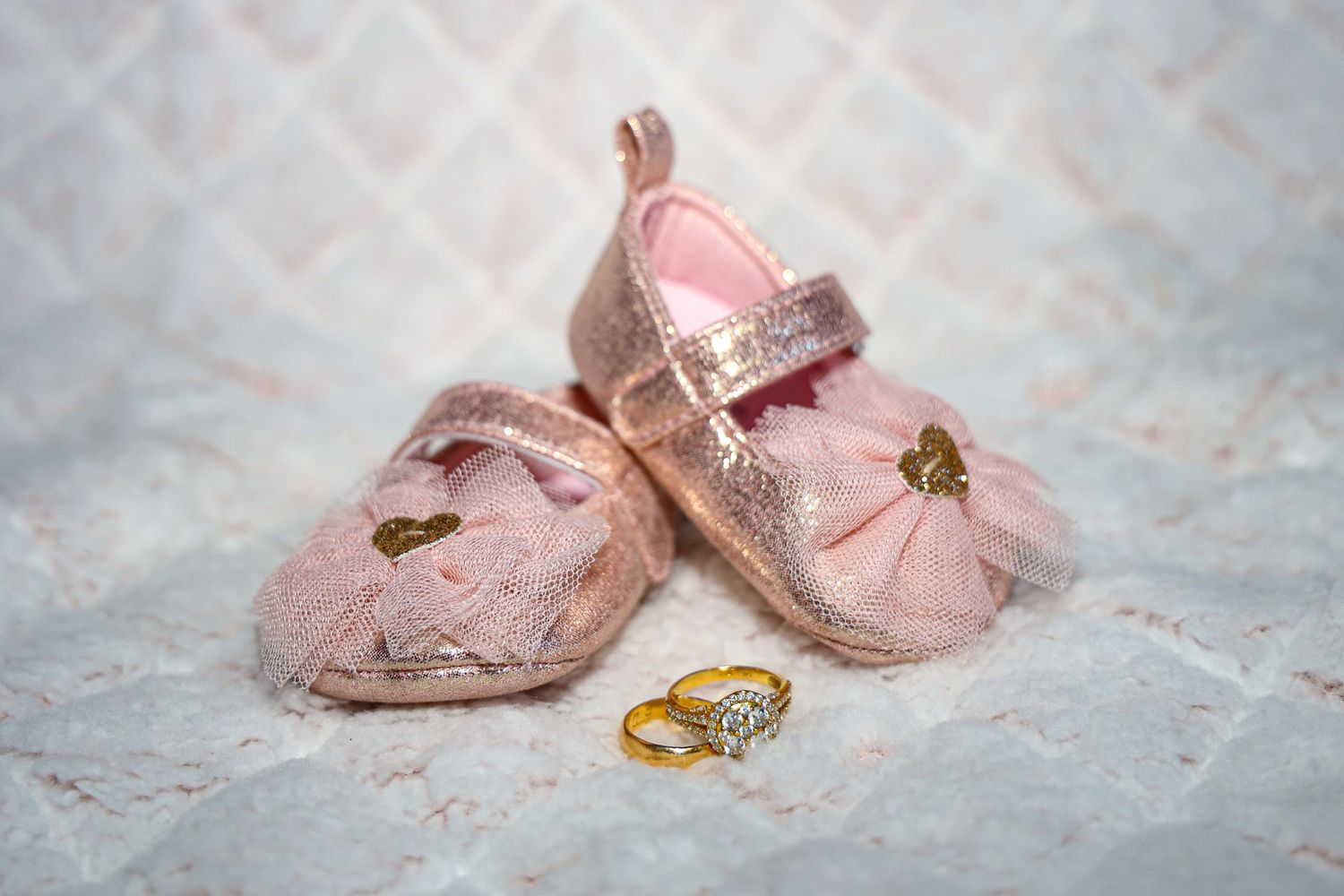 Pink baby shoes with gold butterflies and a matching engagement ring displayed on delicate lace background.