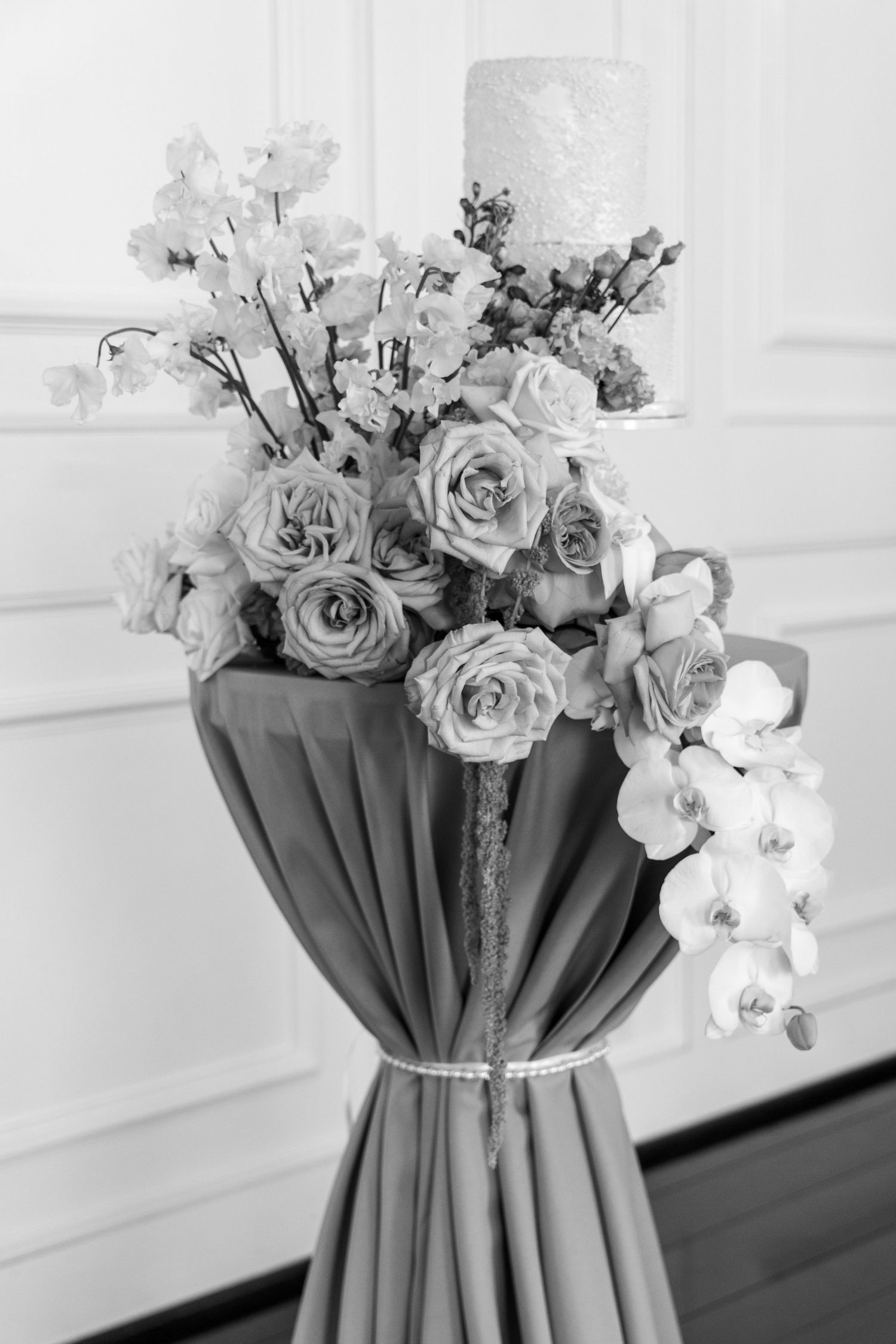 Black and white photo of an elegant floral arrangement with roses and orchids on a gathered fabric stand.
