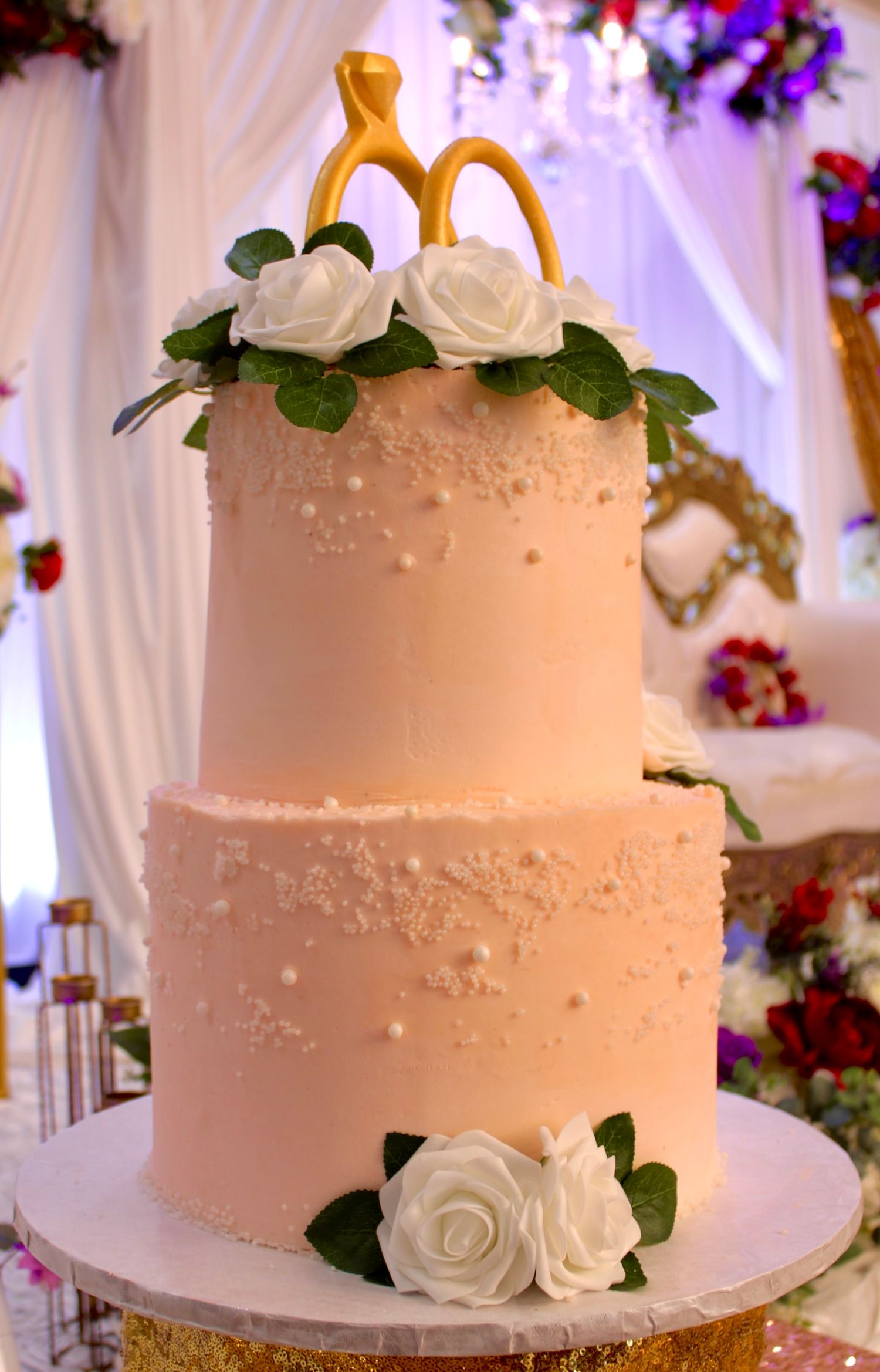 Elegant two-tier white wedding cake decorated with delicate white roses and a golden topper in a romantic reception setting.