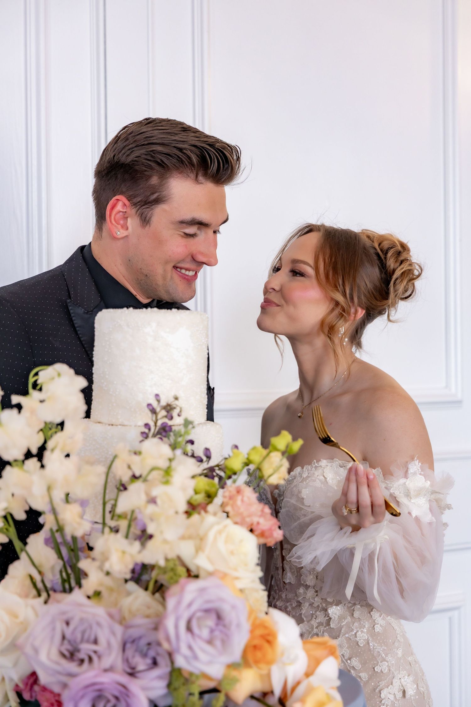 A couple shares an intimate moment beside their white wedding cake adorned with fresh flowers.