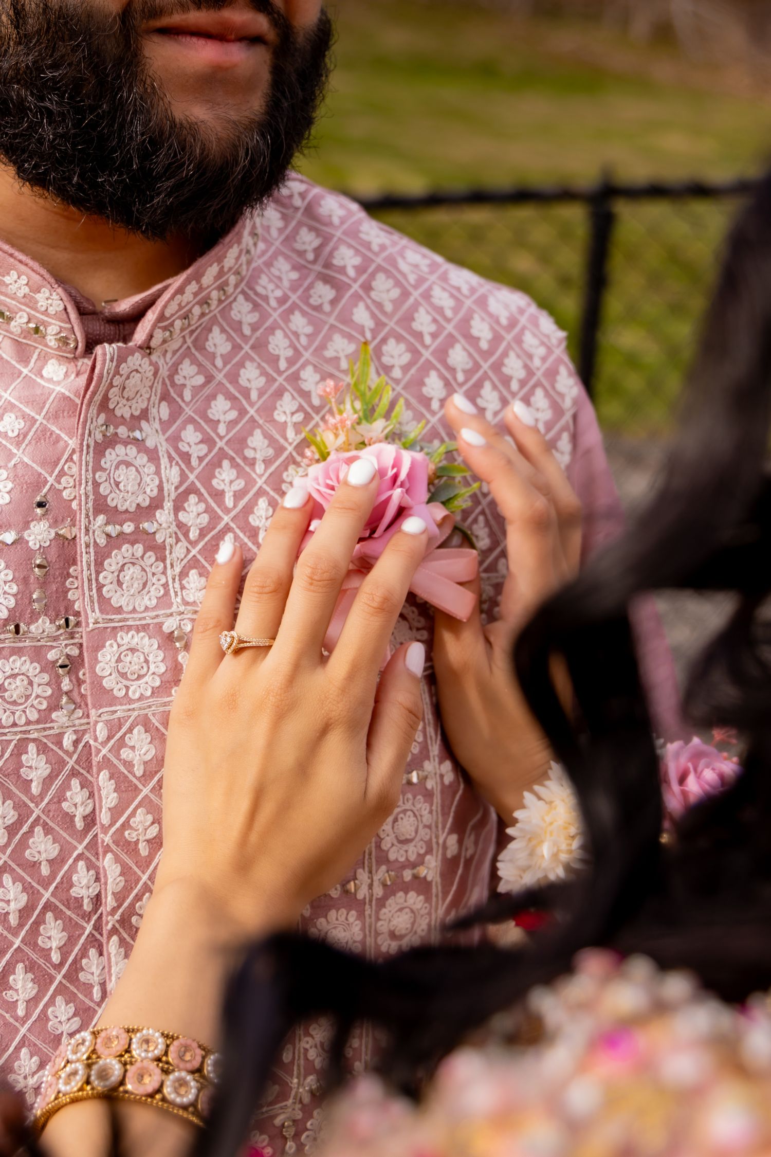 Hands holding delicate pink flowers while wearing a pink patterned shirt and gold bracelet against a blurred outdoor background.
