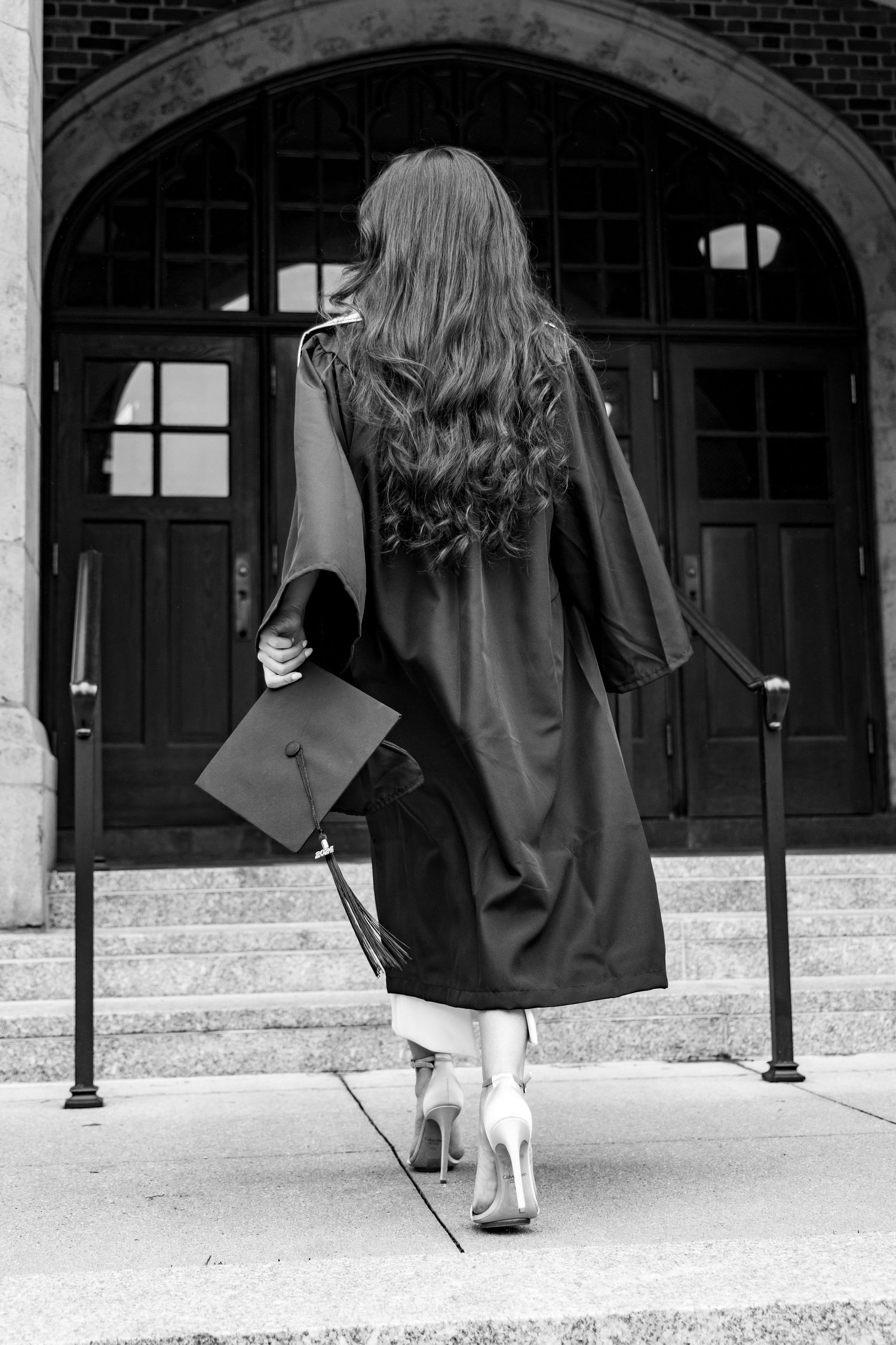 Black and white graduation photo showing graduate in cap and gown from behind ascending stone steps at building entrance.