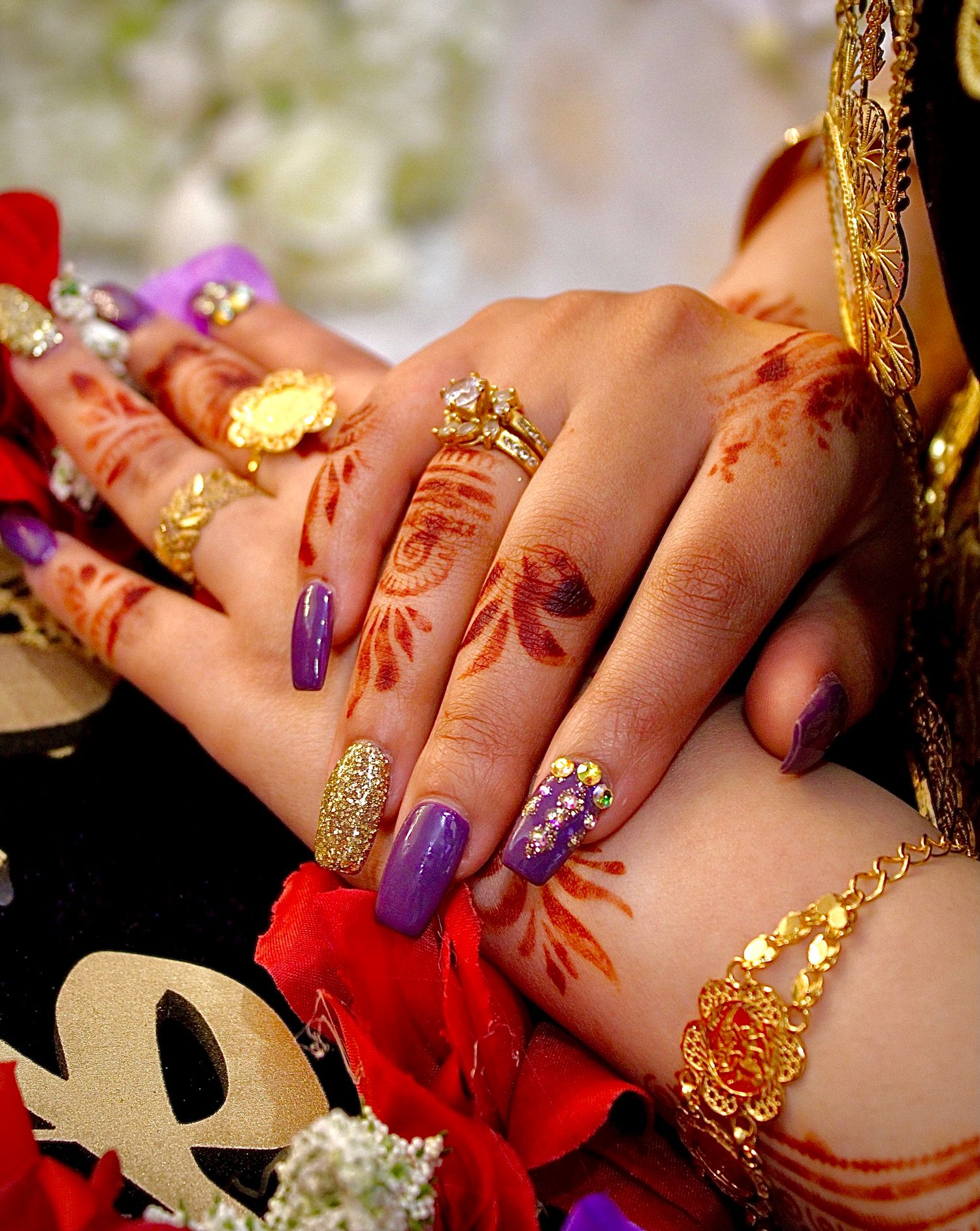 Ornate henna-decorated hands with gold jewelry wearing purple and glitter nail art beside a red flower.