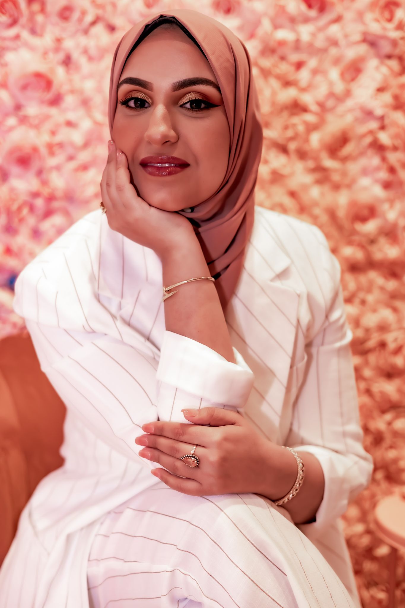 Elegant woman in white blazer and mauve hijab poses against a pink floral background with hand resting on face.