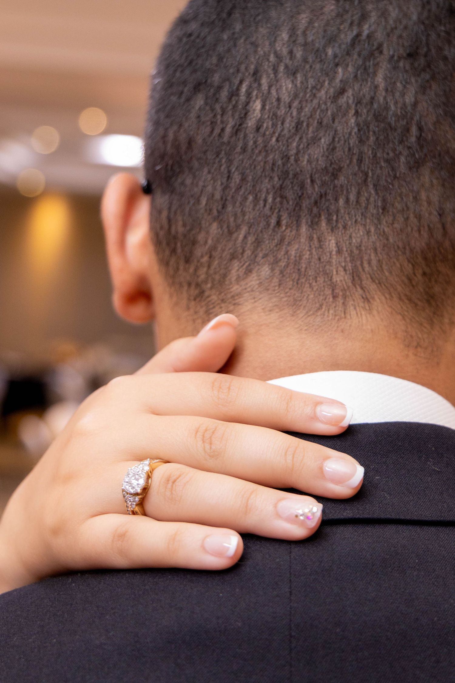 A hand with a sparkling diamond ring embraces someone wearing a dark suit jacket in an intimate moment.