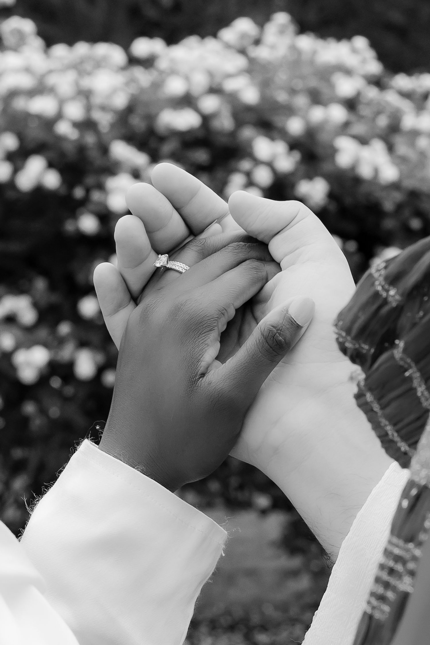 Two hands clasped together wearing white sleeves with white flowers in the background, captured in black and white. | PHL, PA Wedding