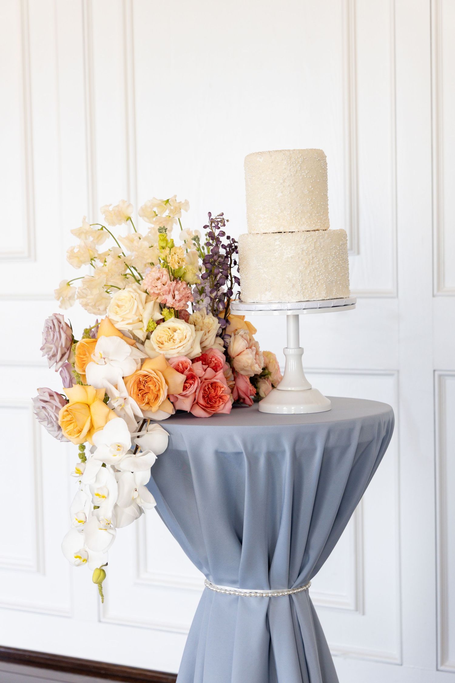 Elegant two-tier white textured wedding cake on pedestal with cascading peach and cream floral arrangement on gray tablecloth.