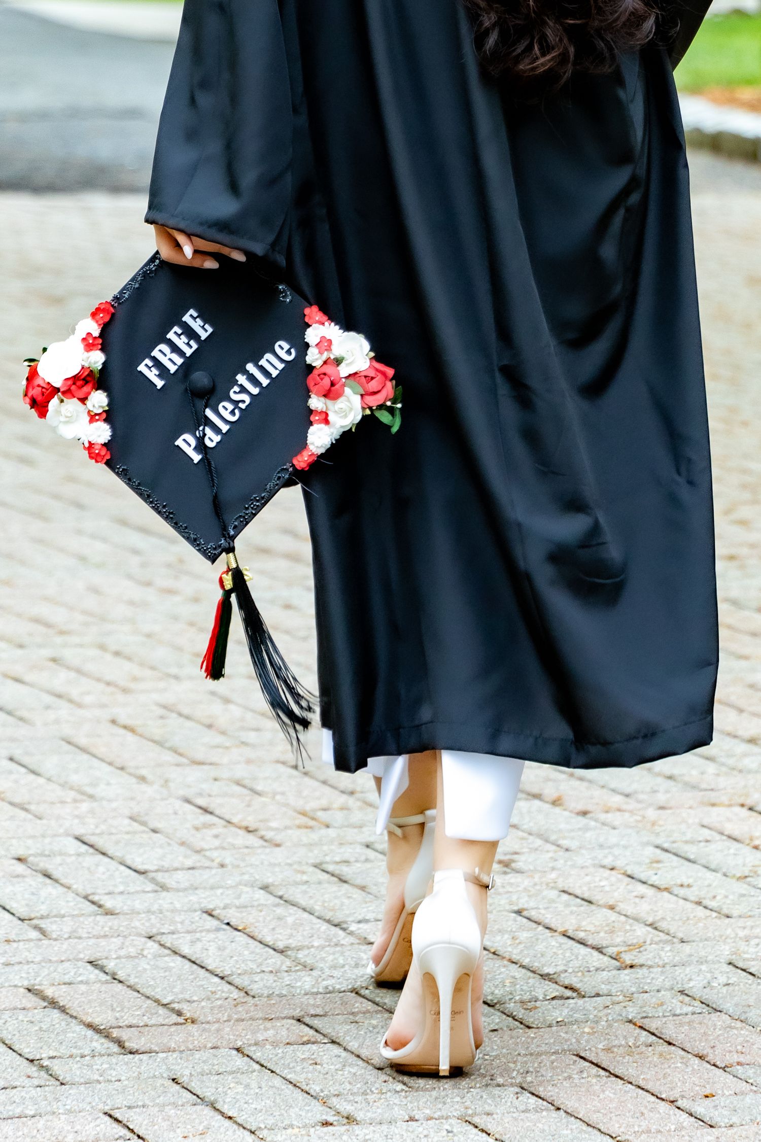 Graduation cap decorated with red flowers and the words 'Fire Palestine' worn with a black gown and beige heels.