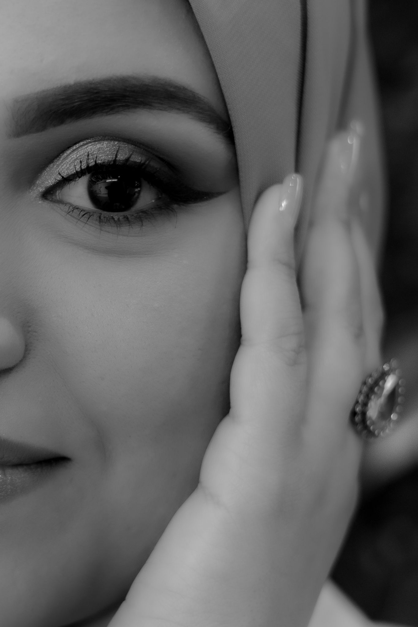 Close-up black and white portrait showing an eye with dramatic makeup and a hand wearing a ring.
