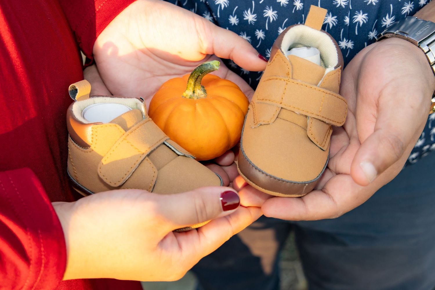 Hands hold tiny brown autumn booties alongside a small decorative pumpkin against a dark background.