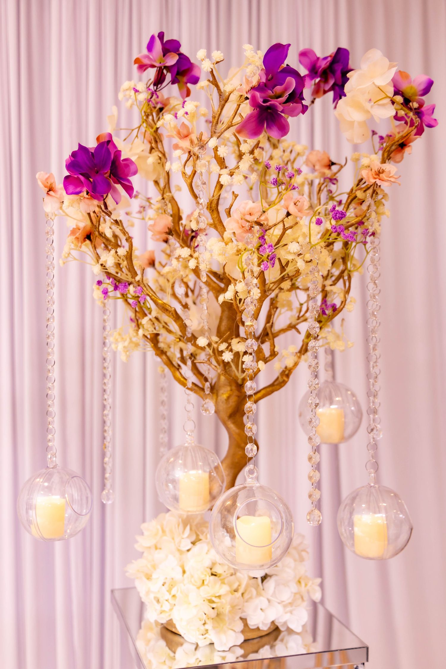 Elegant centerpiece with purple orchids on gold branches with hanging glass candle holders against a white backdrop.