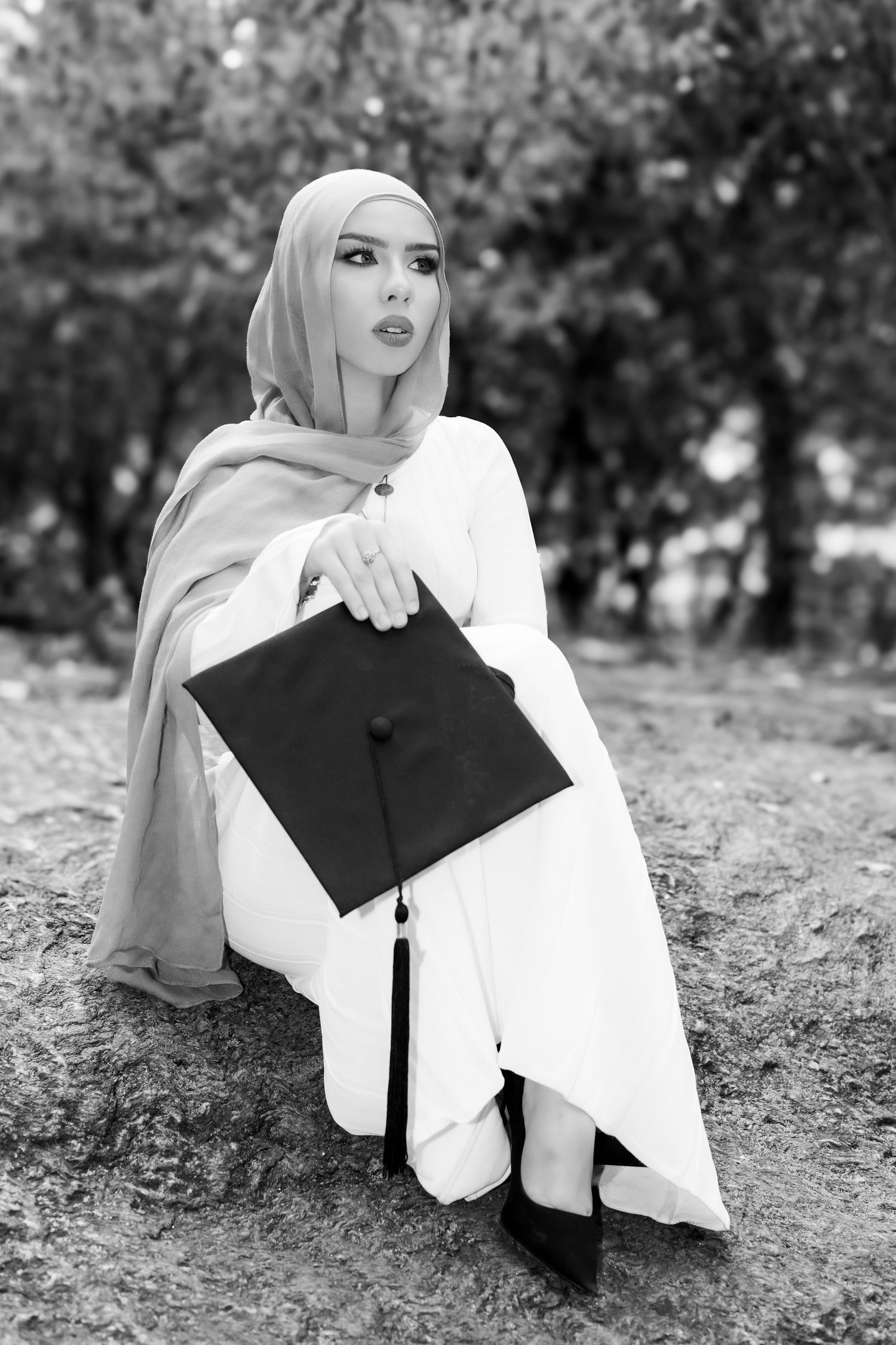 Black and white portrait of graduate in hijab and gown holding graduation cap while sitting outdoors.