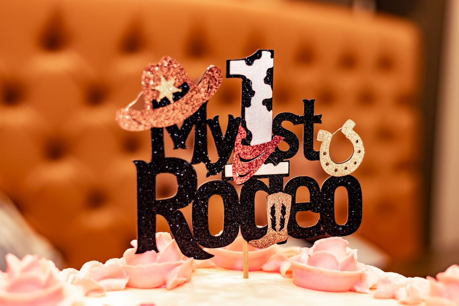 Black 'My 1st Rodeo' cake topper with cowboy silhouette decorated with pink accents on tufted background.