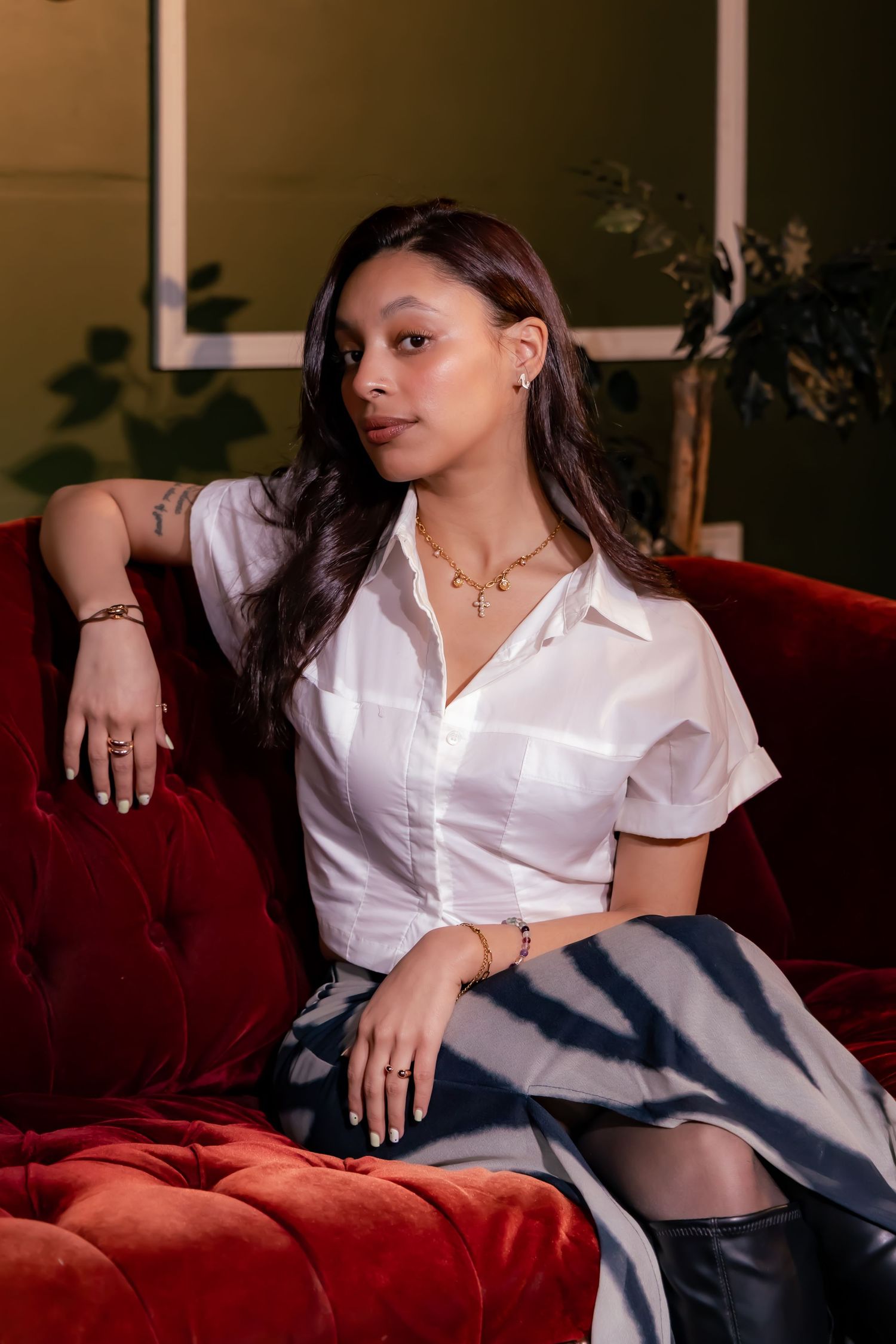 Someone wearing a white button-down shirt and dark skirt poses elegantly on a plush red velvet couch in a dimly lit room.