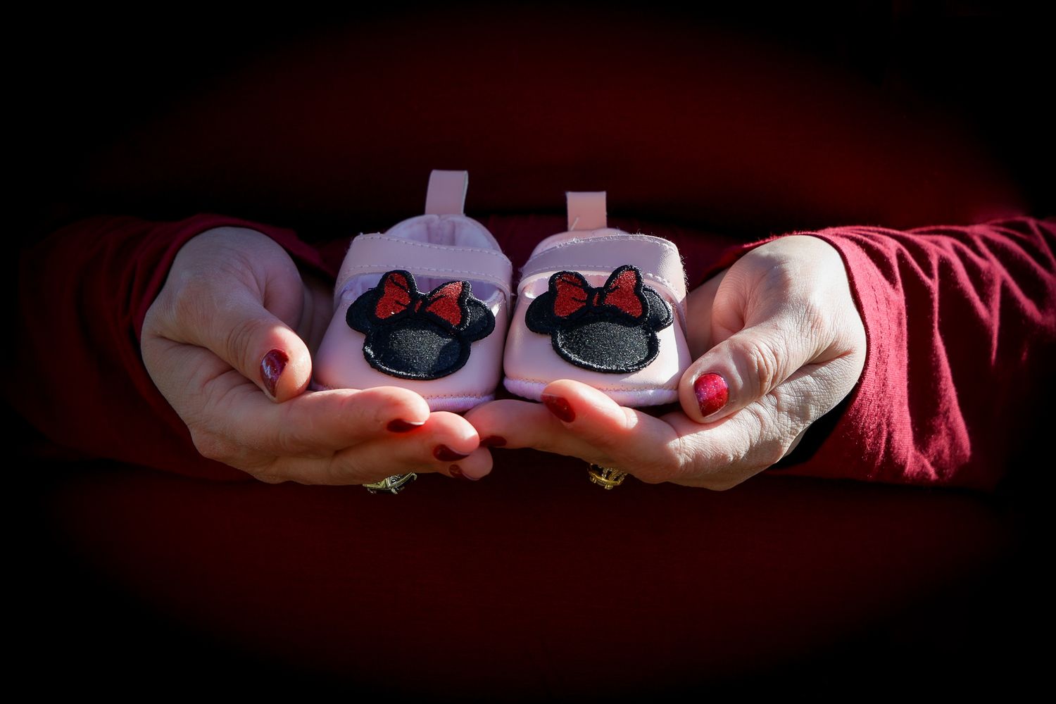 Hands holding two white baby booties decorated with Minnie Mouse bows against a dark background.