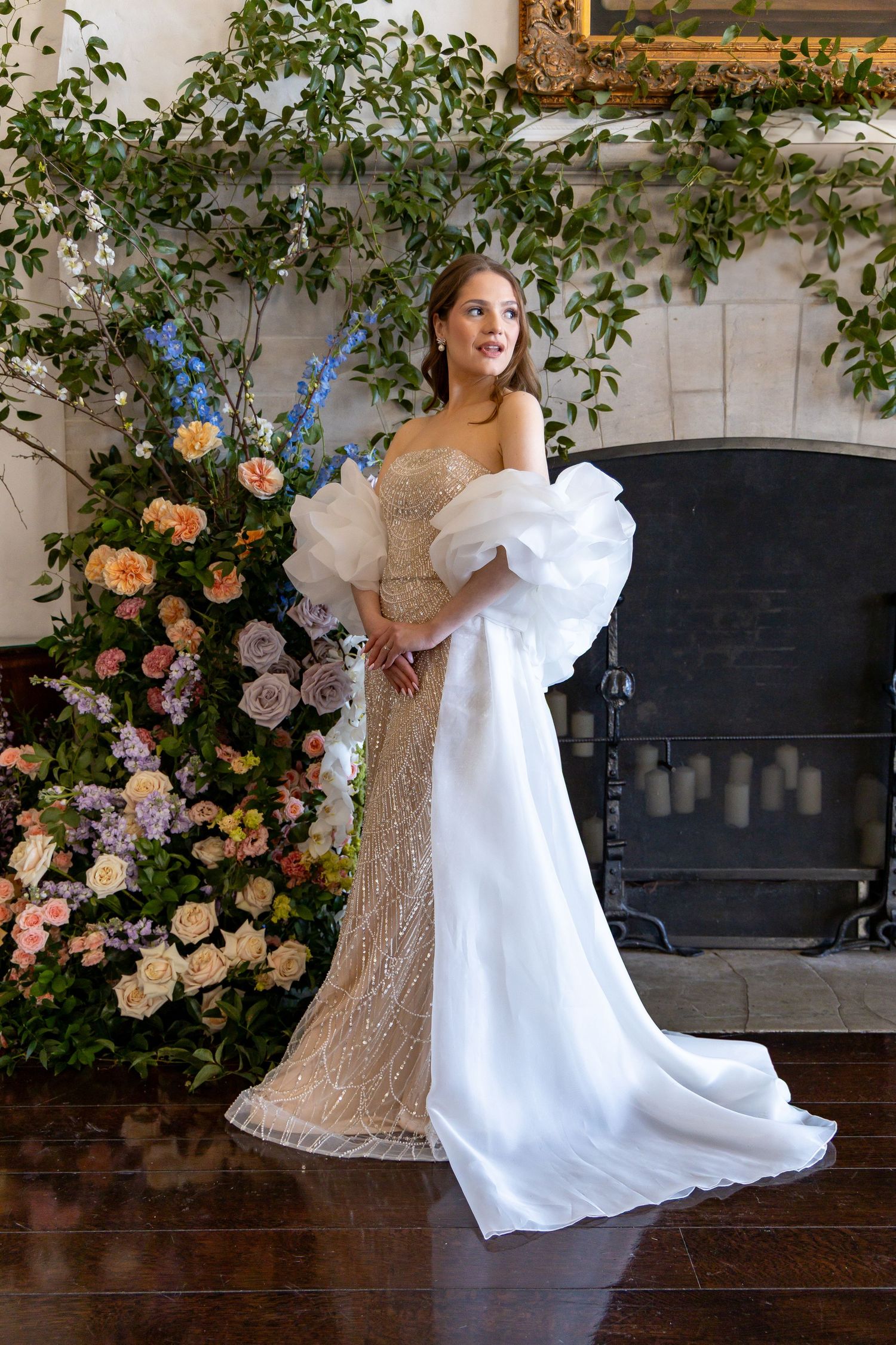 Elegant bridal portrait featuring a lace gown with dramatic white overlay sleeves against a floral-adorned fireplace.