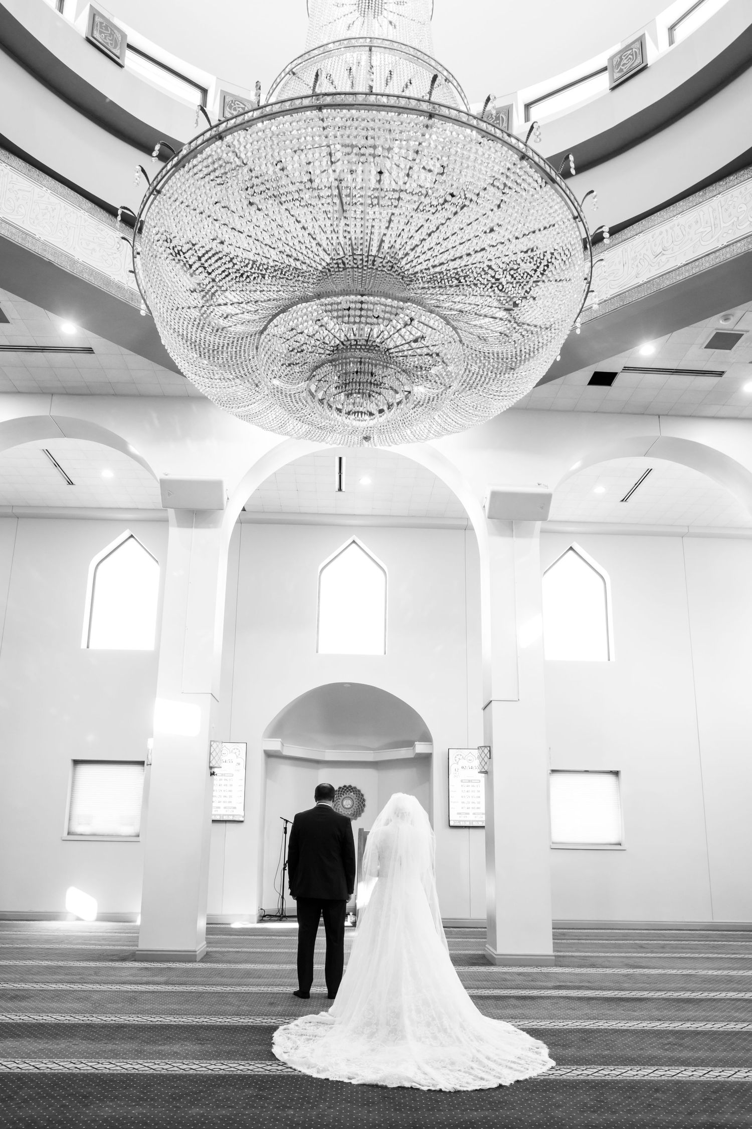 Black and white photo of bride and groom standing beneath grand chandelier in elegant mosque interior.