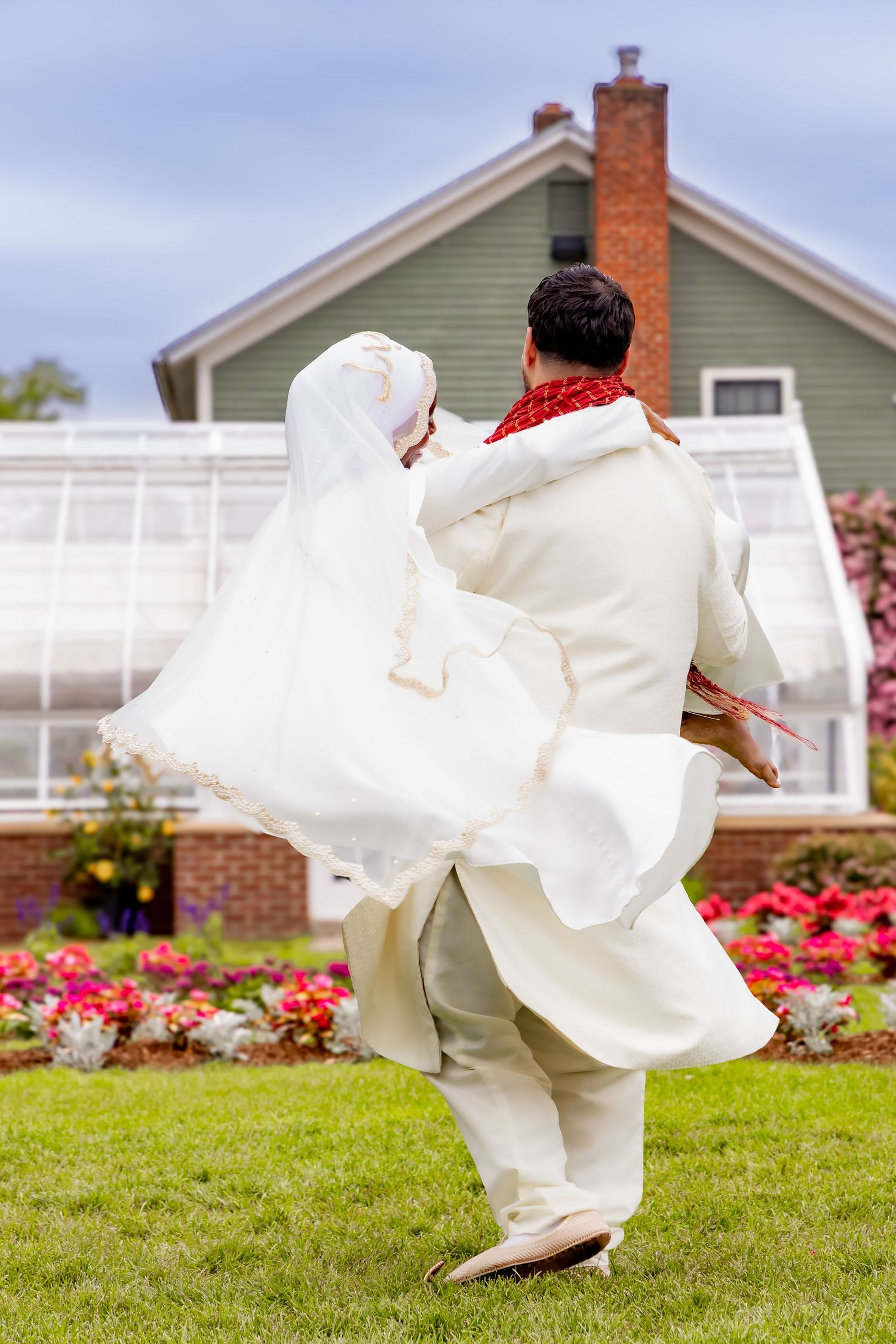 A couple in white wedding attire dances on a lawn with a green house and red flowers in the background. | PHL, PA Wedding Photographer