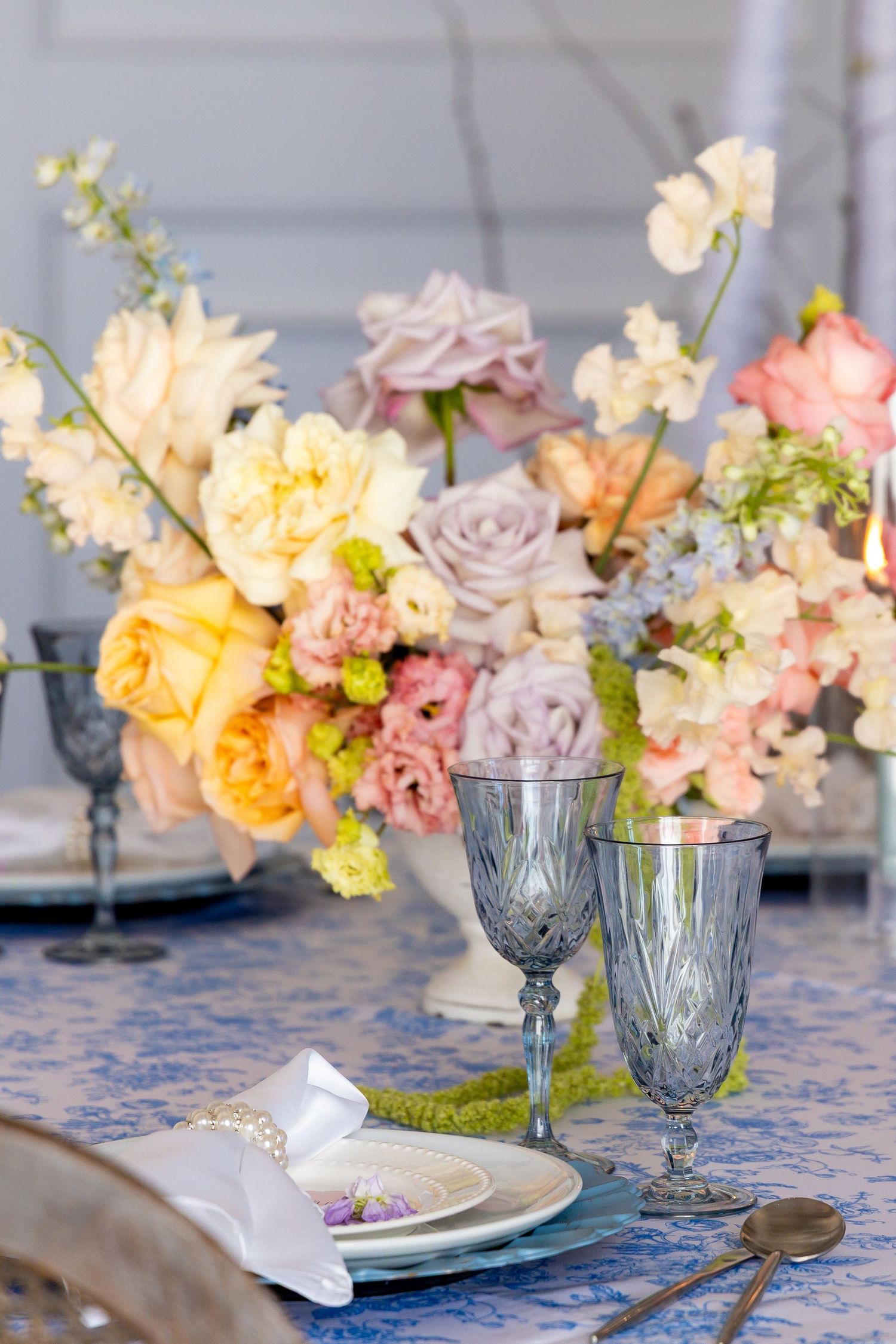 Elegant table setting with vintage blue glassware and pastel floral centerpiece featuring roses and delicate blooms on blue cloth.