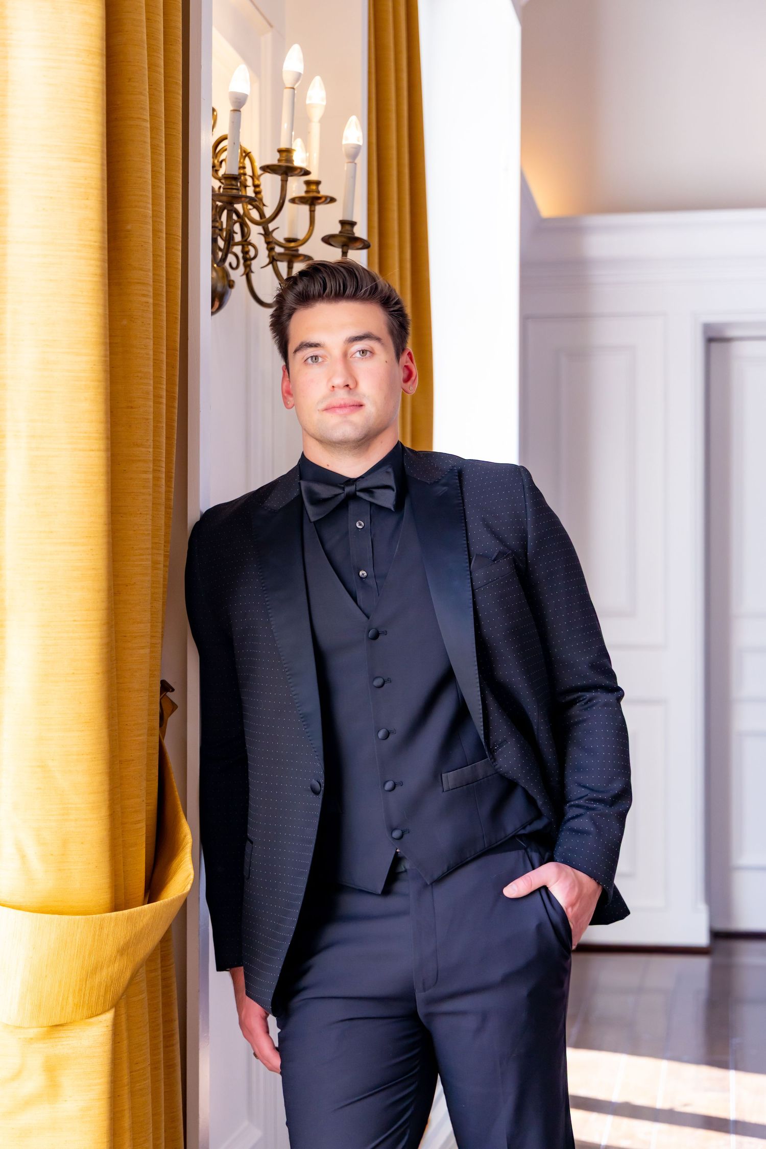 Elegant groom in black tuxedo poses against gold curtain and ornate wall sconce.