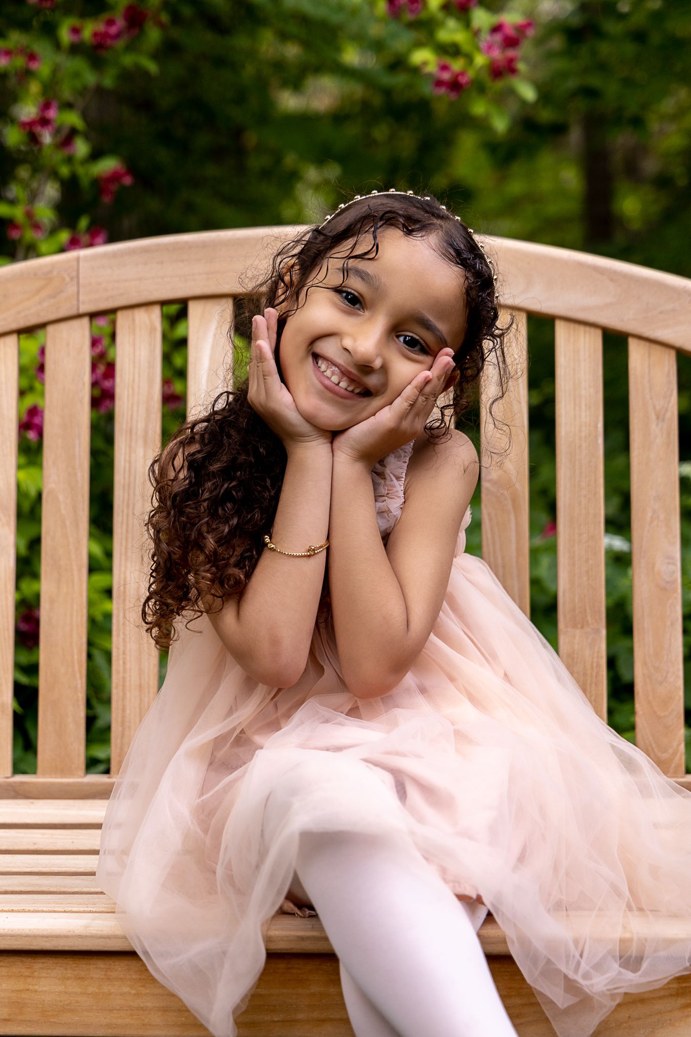 Child in pink tulle dress and white tights poses sweetly on wooden garden bench with flowering bushes in background.