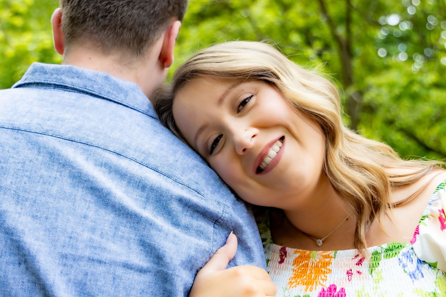 A romantic couple embraces outdoors on a sunny day with lush green foliage in the background.