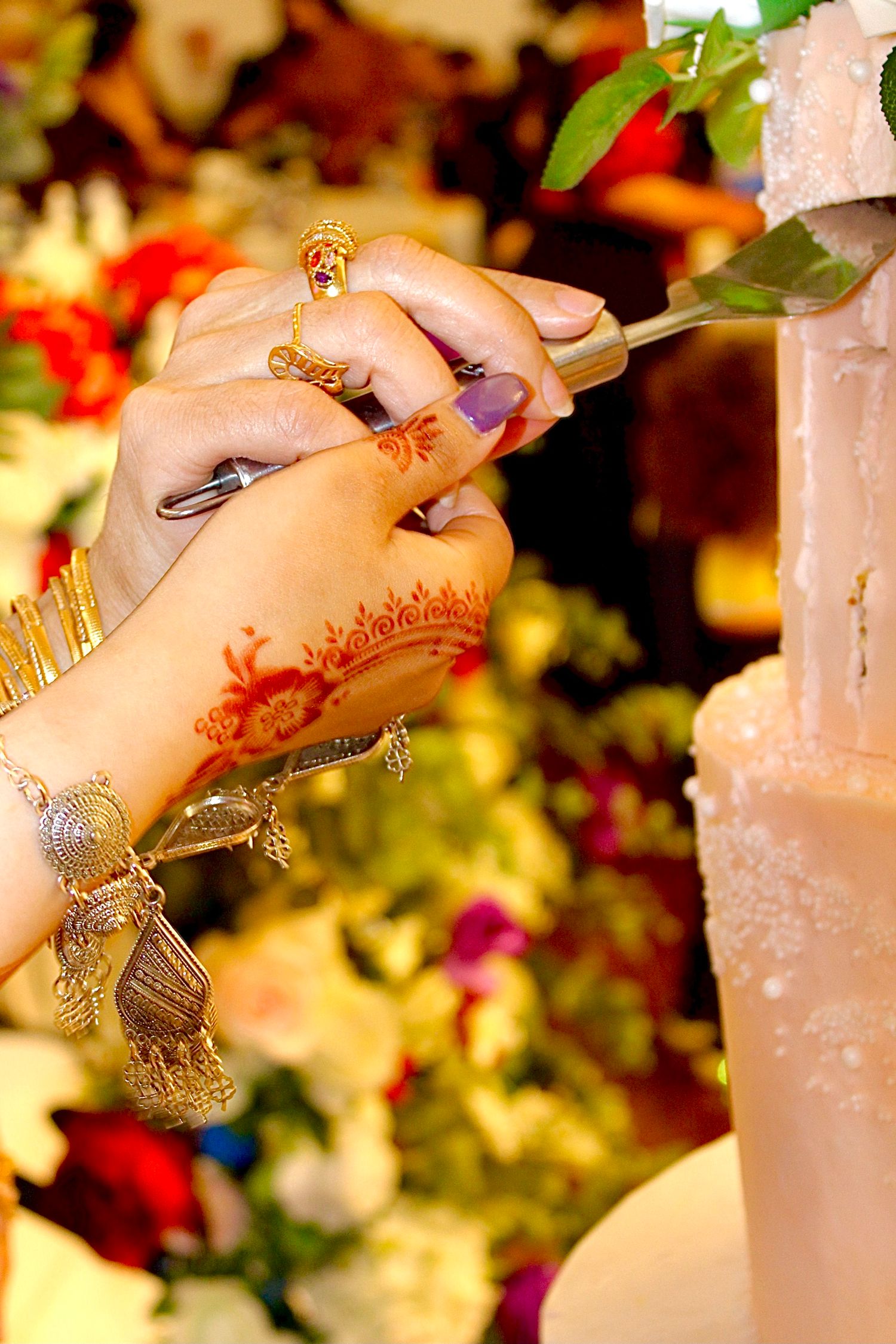 Hands adorned with henna and gold jewelry cut into a wedding cake against a blurred floral background.