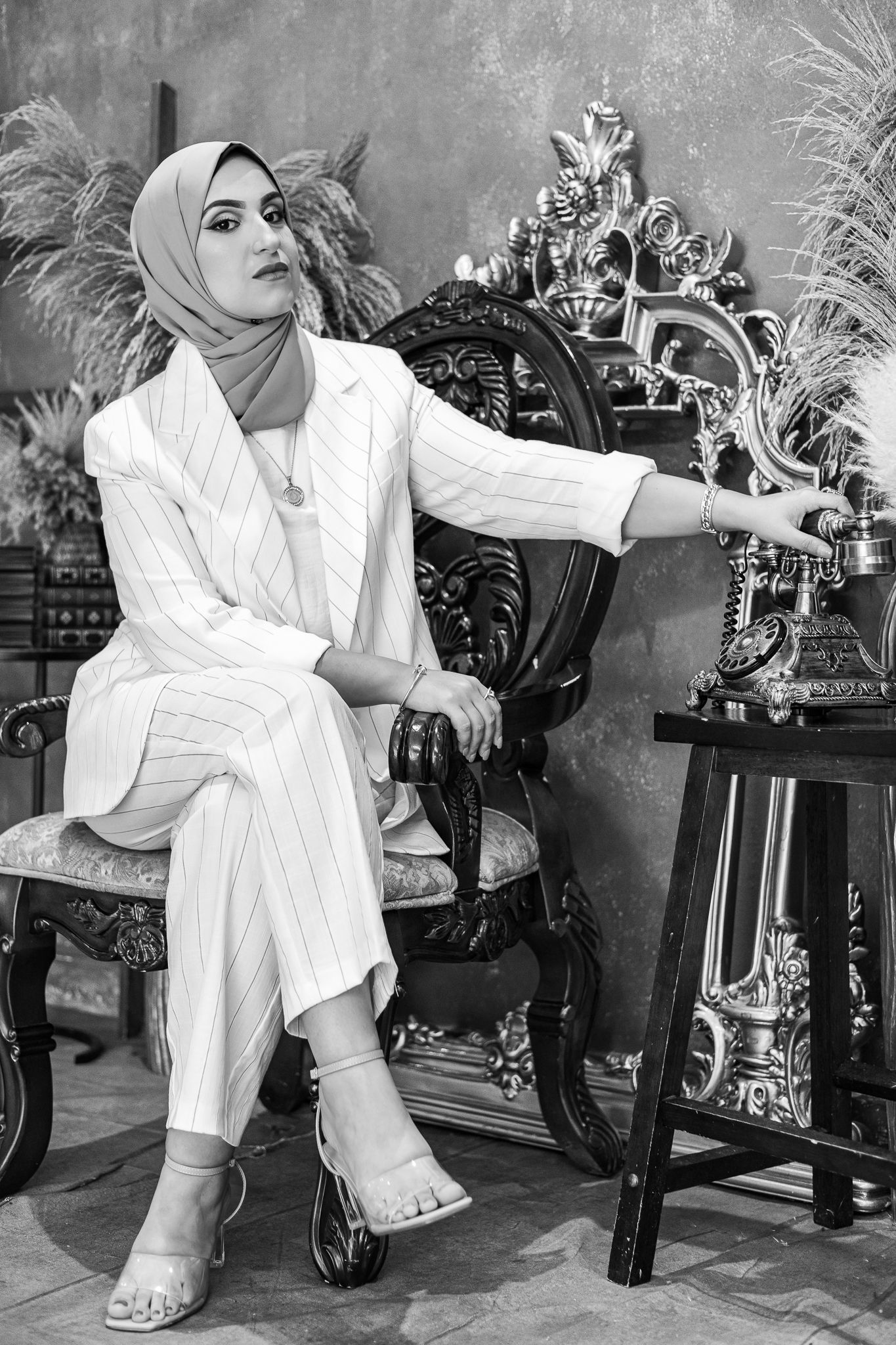 Black and white portrait of someone in a white pantsuit and hijab sitting elegantly in an ornate vintage chair.