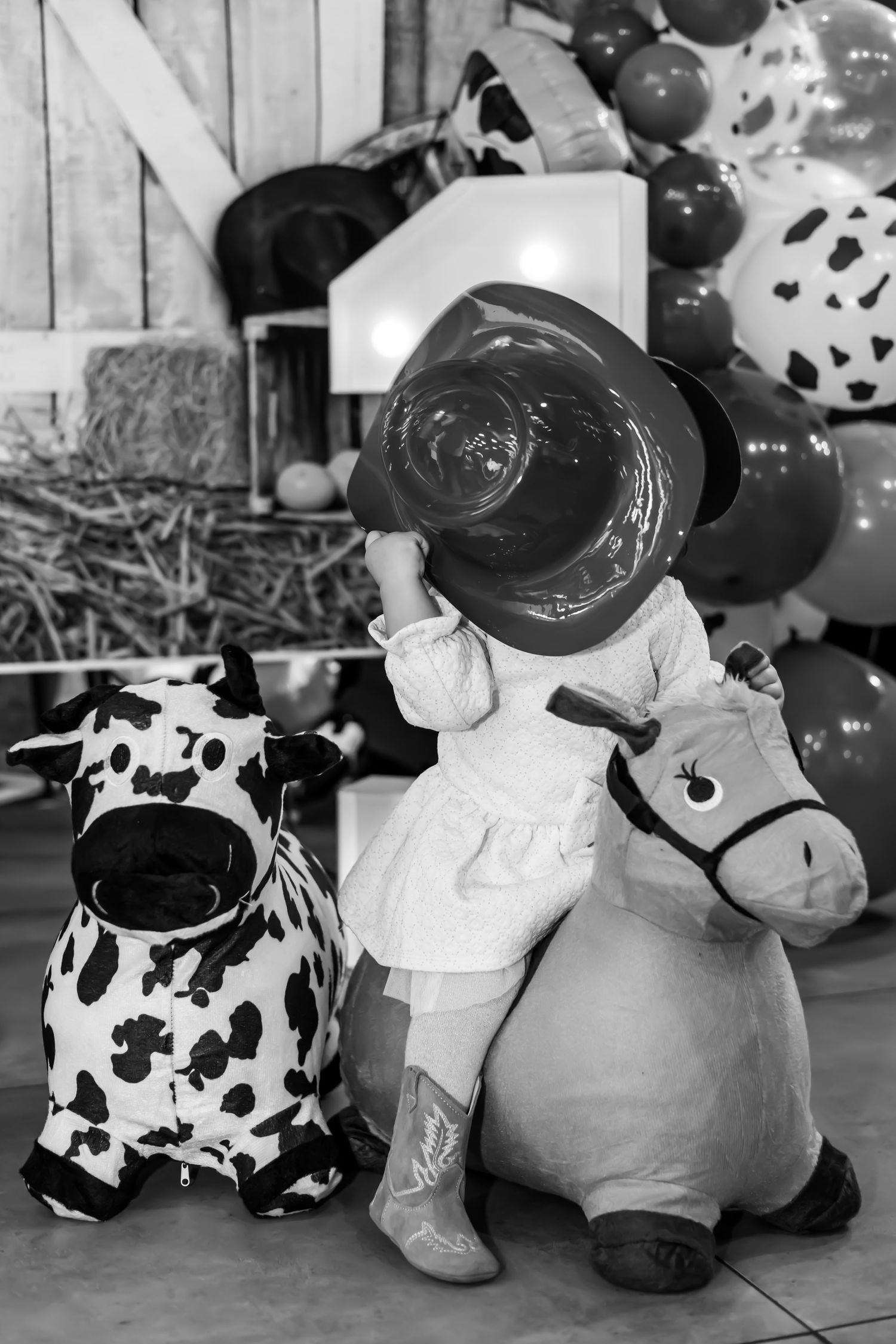 A black and white photo showing a stuffed toy cow and rocking horse with cowboy-themed party decorations on a farm.