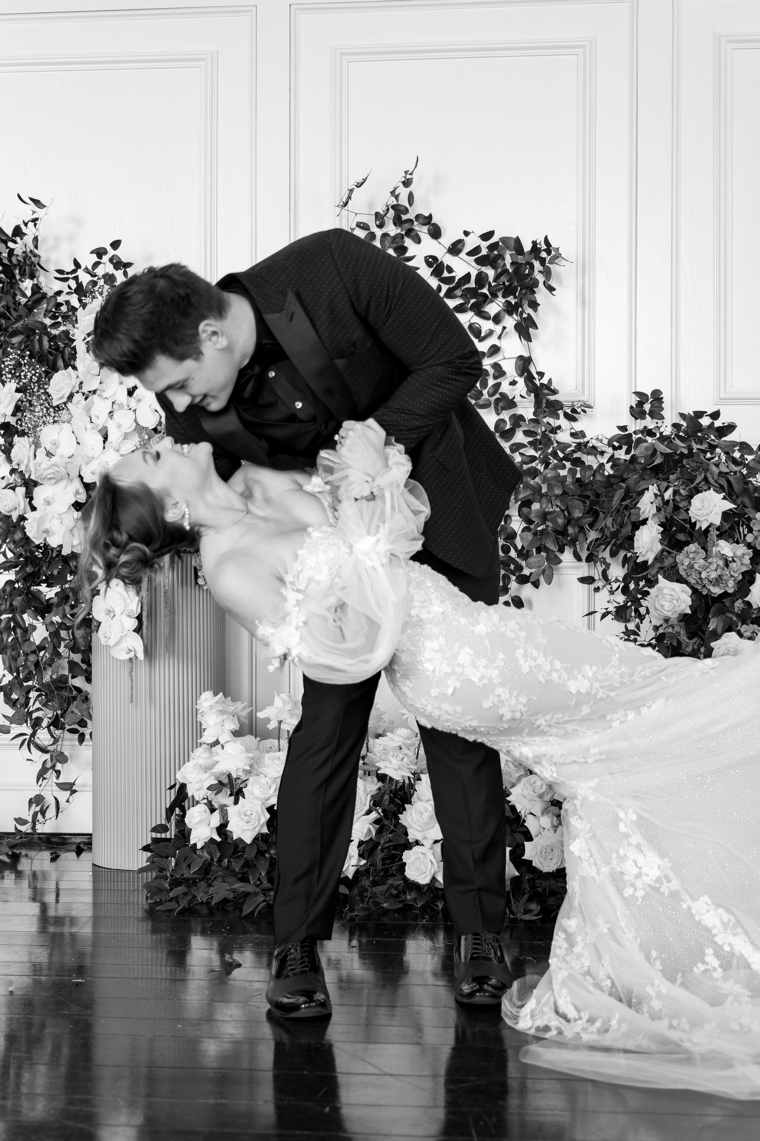 Romantic black and white photo of wedding couple in dramatic dip pose with floral backdrop.