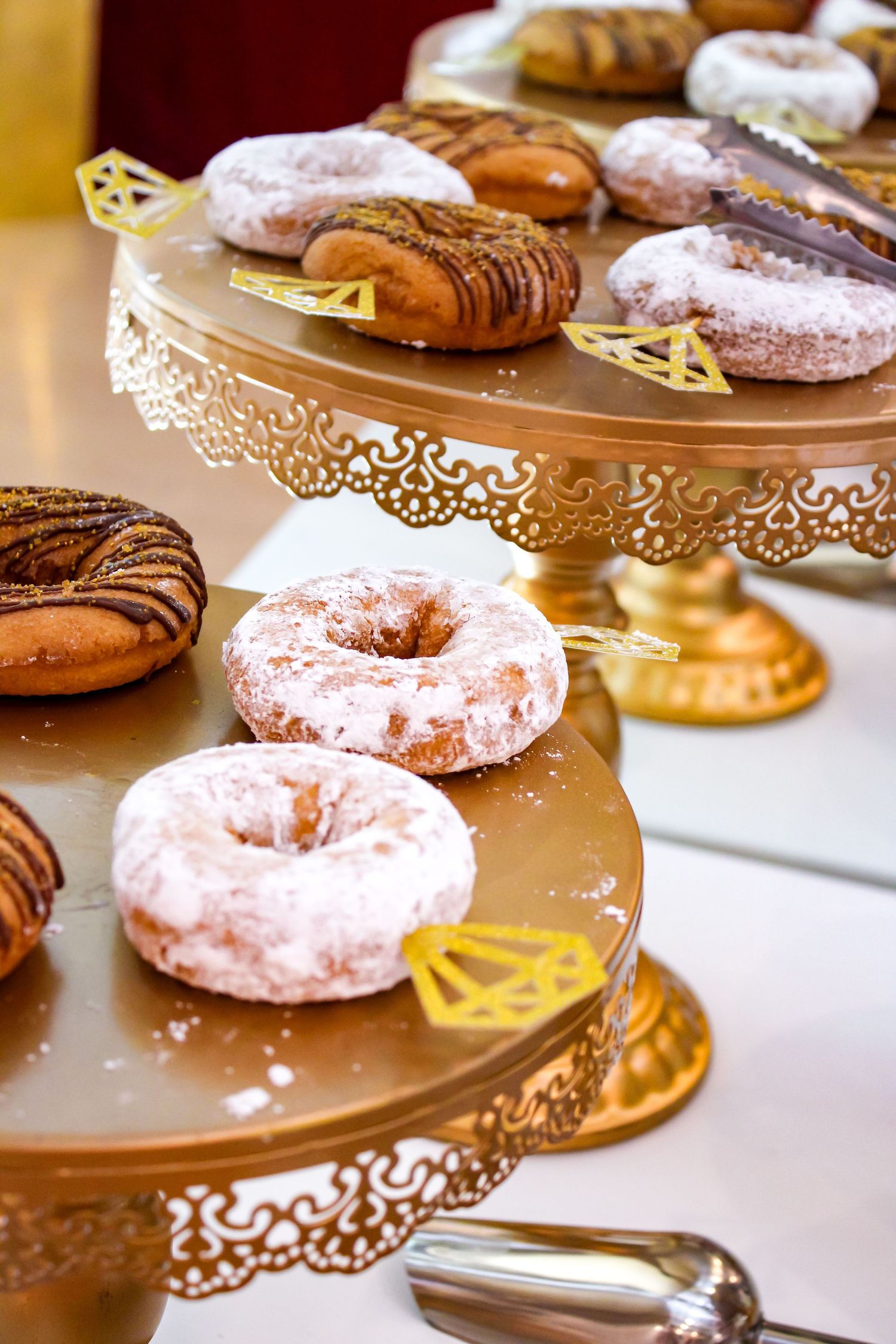 Pink frosted and chocolate glazed donuts displayed on elegant gold tiered dessert stands with decorative lace edges.