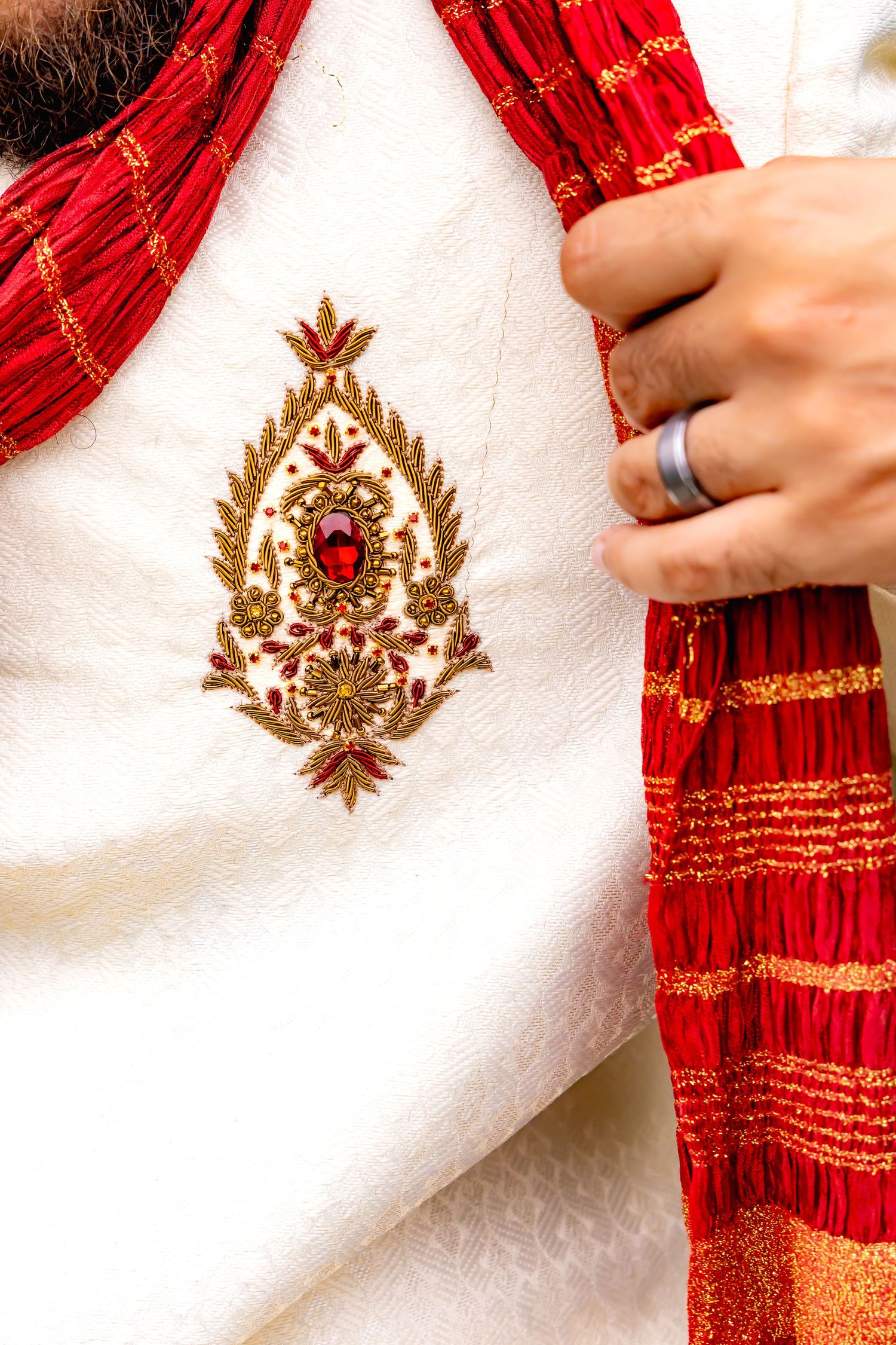 Ornate gold embroidered design with red gemstone on white fabric next to red and gold traditional scarf. | PHL, PA Wedding Photographer