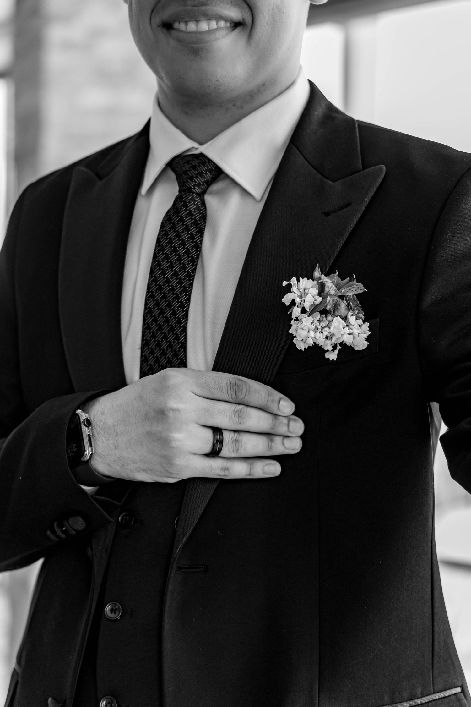 Black and white photo showing a groom in formal suit with boutonniere and tie placing hand with wedding ring.